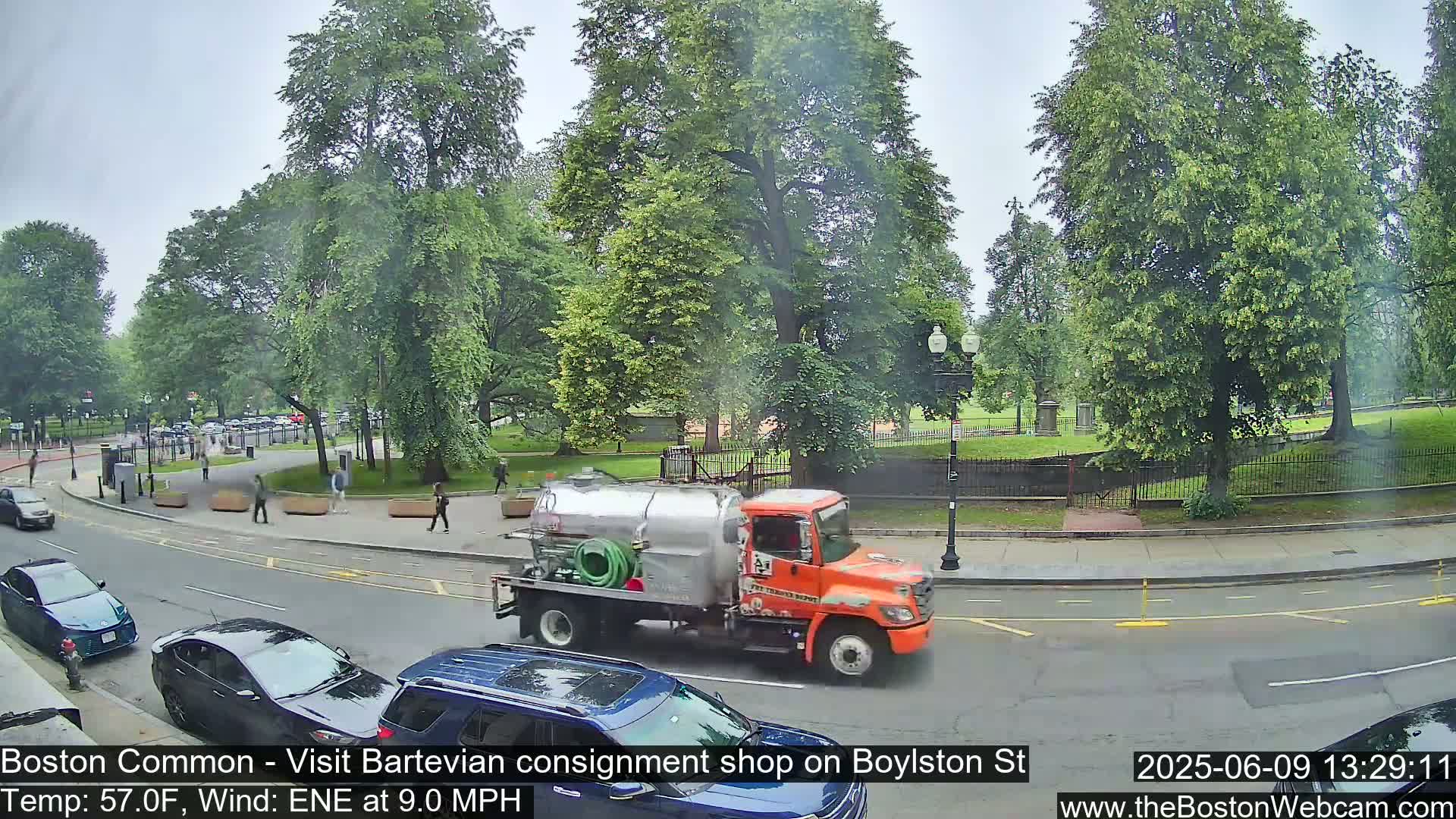 A city street scene on an overcast day shows a parked tanker truck next to several cars, with pedestrians walking along a sidewalk adjacent to a grassy park.