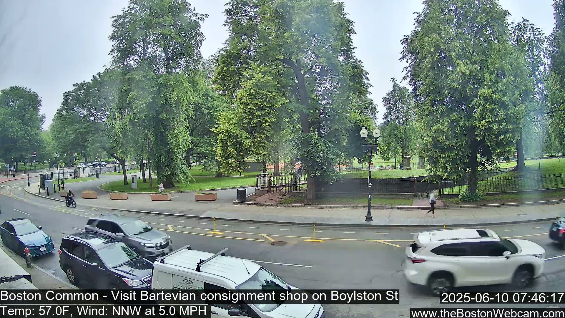 A city street scene on an overcast day shows several cars driving past a park with lush green trees and people walking, along with a person on a bicycle.