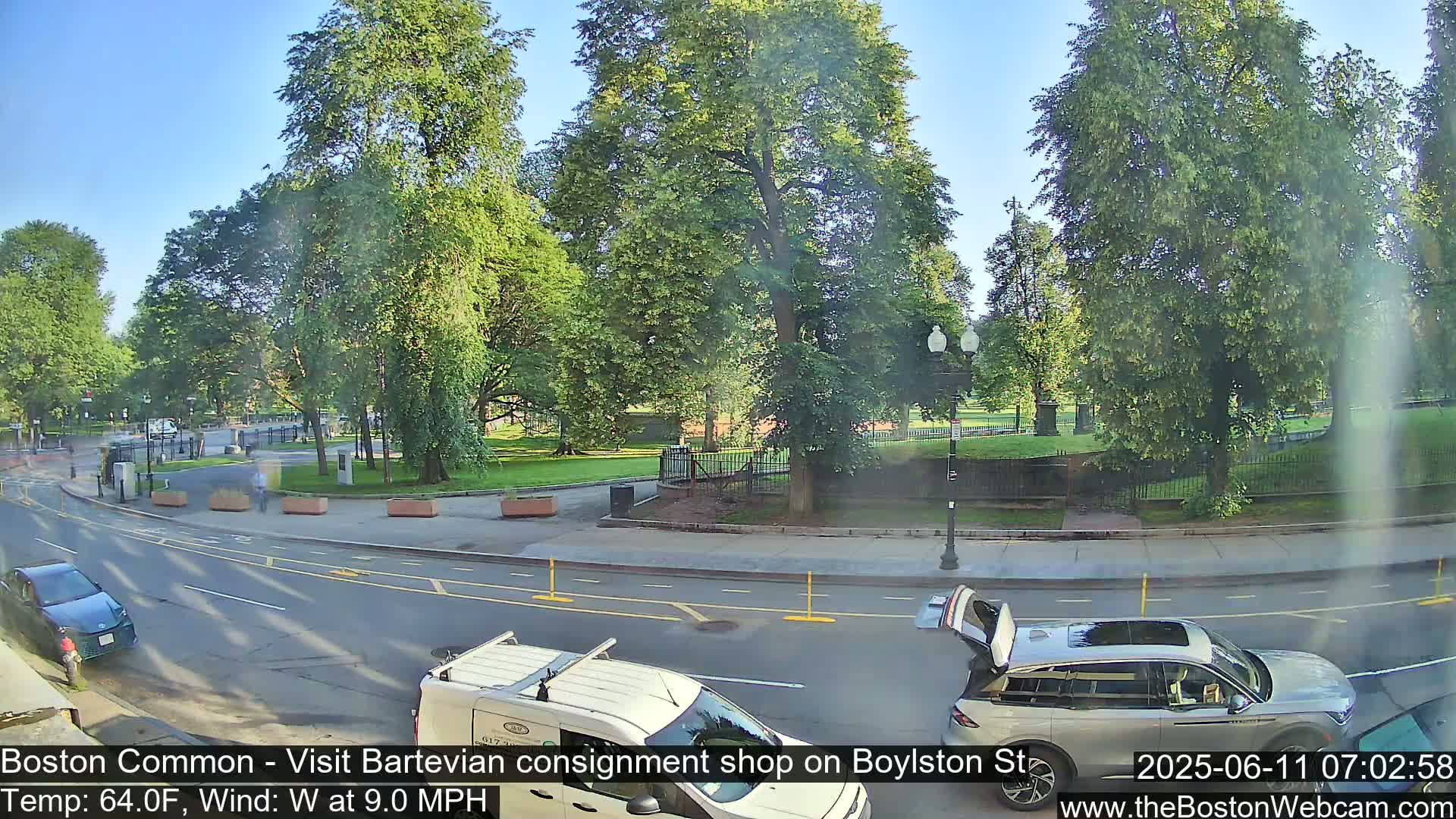 A sunny, partly cloudy day shows a street scene with cars and a van near a park filled with lush green trees.