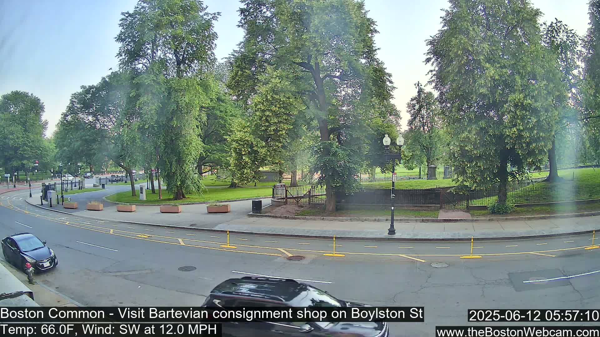A partly sunny day shows a curved road next to a park with lush green trees and manicured lawns, with cars driving on the road.