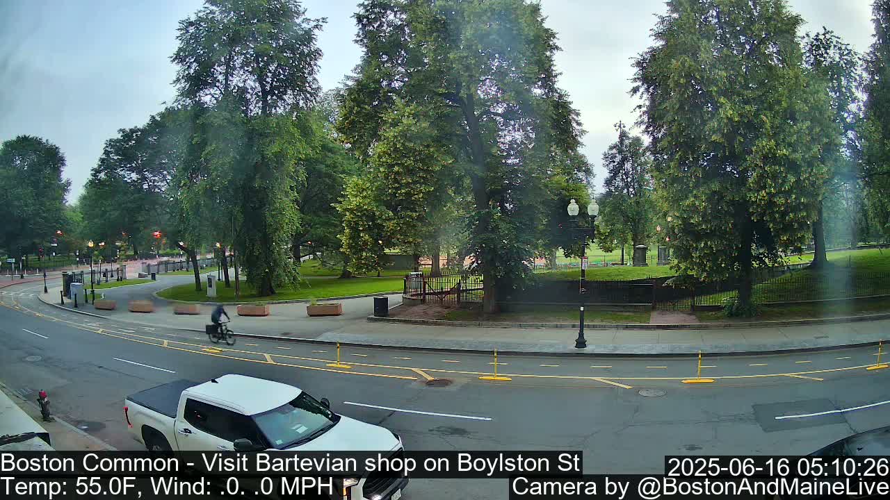 A mostly cloudy day shows a street scene with a white pickup truck, a bicyclist, and lush green trees surrounding a park.
