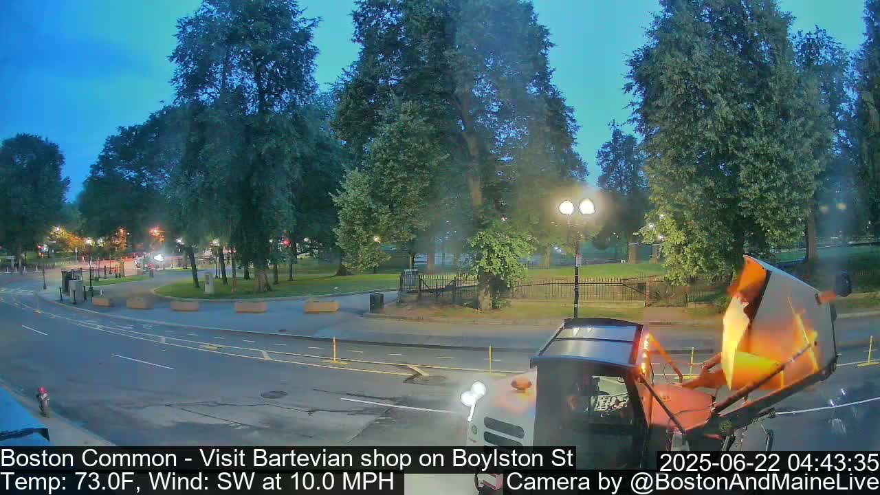 A street scene at dusk shows a partially visible garbage truck near a park with numerous trees under a clear, twilight sky.