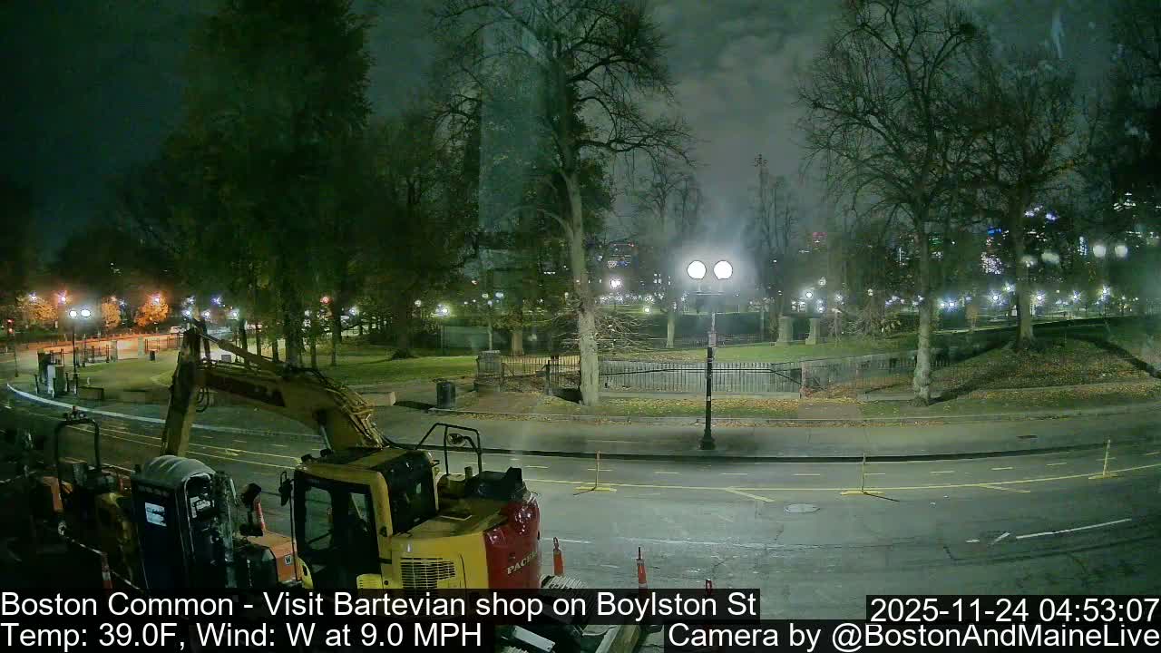 An excavator and other construction equipment are parked on a wet street next to a park illuminated by streetlights under a misty night sky.