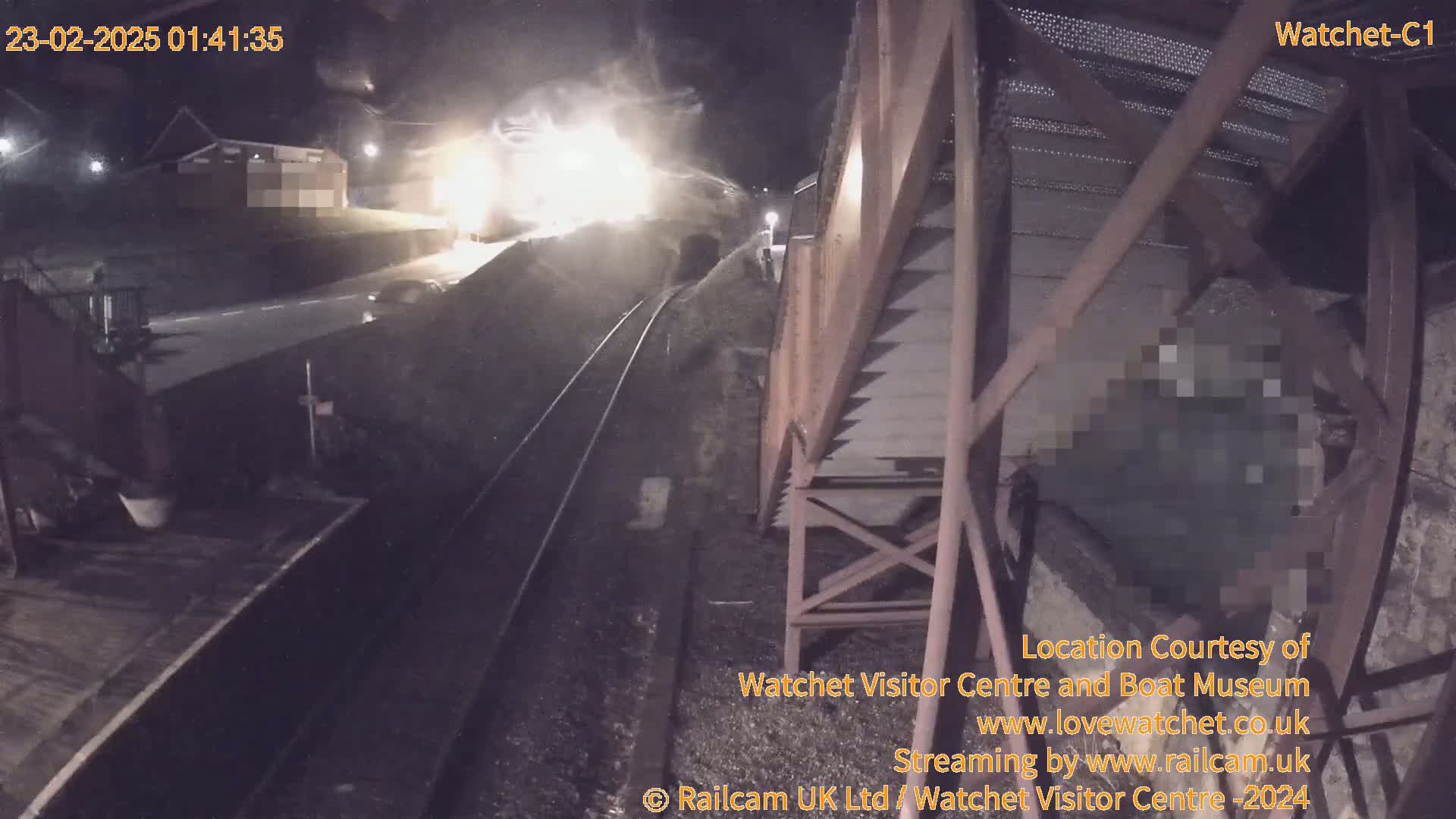 A nighttime view of a train passing by a railway track next to a metal structure, under dark, possibly overcast conditions.