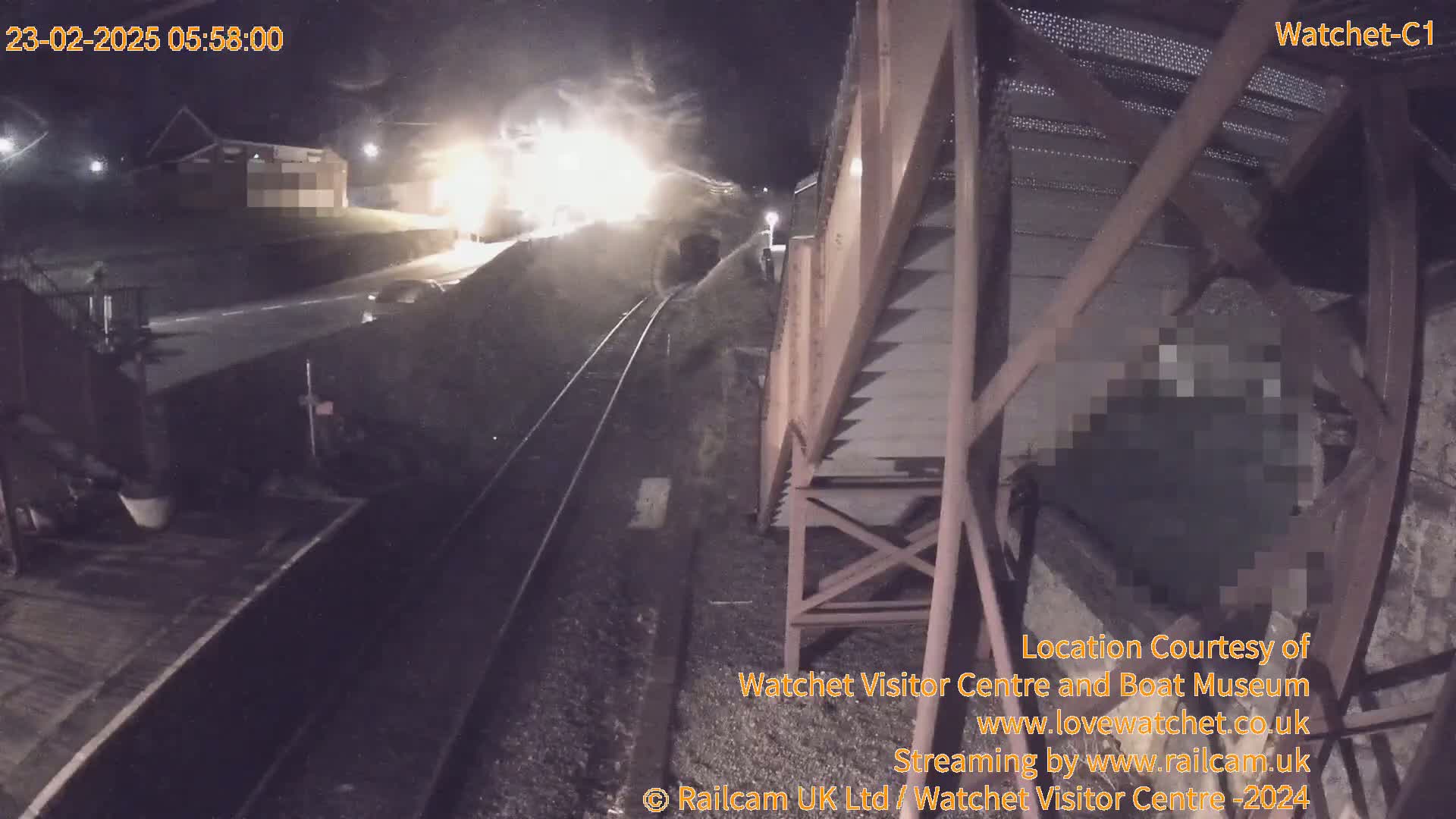 A nighttime, low-angle view shows a train approaching a tunnel under a metal structure, with bright lights illuminating the scene.