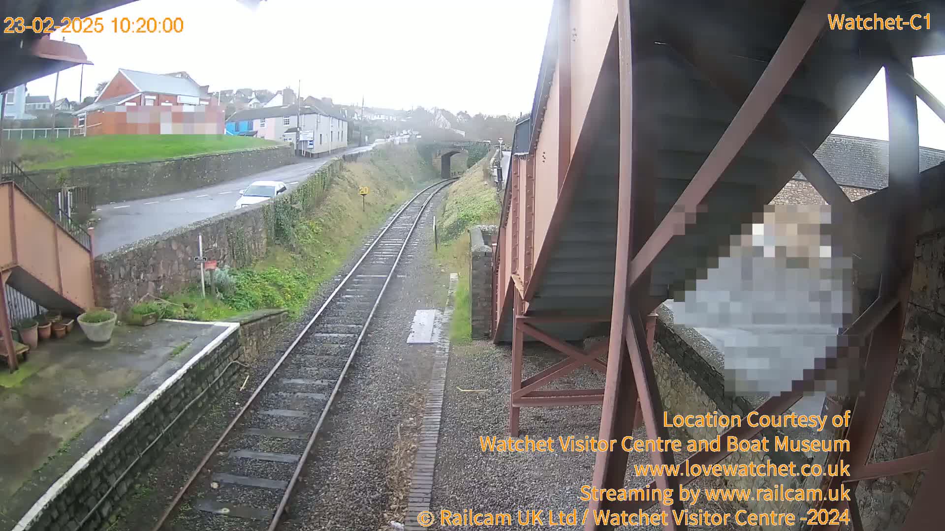 A view from under a railway bridge on an overcast day shows train tracks, a road with a car, and a small town in the distance.