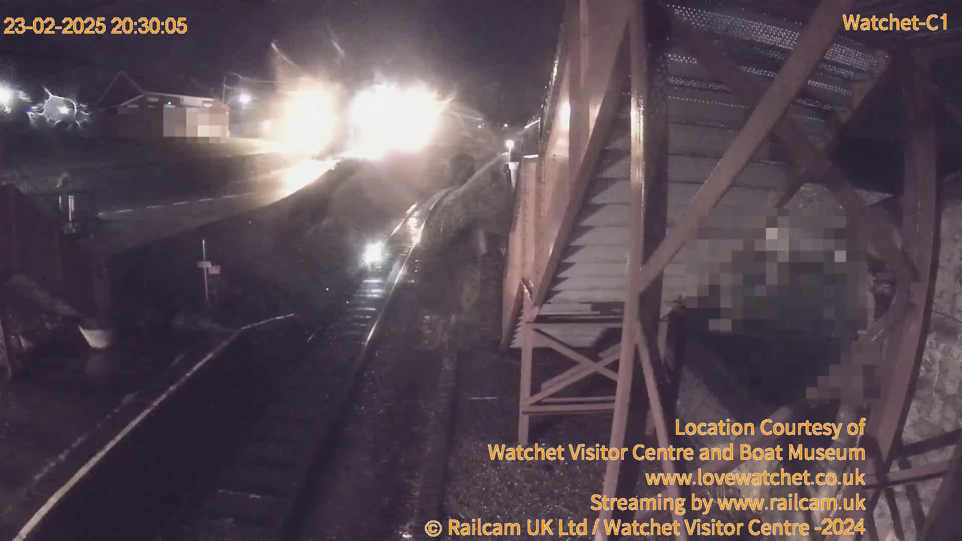 A nighttime, rainy scene shows a train passing under a metal structure near a building.