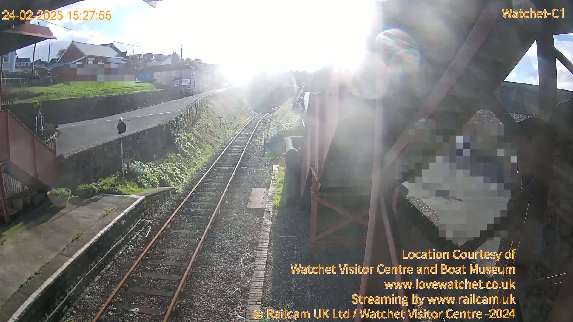 A sunny view from a railway bridge shows train tracks, a person walking on a road next to the tracks, and buildings in the distance.
