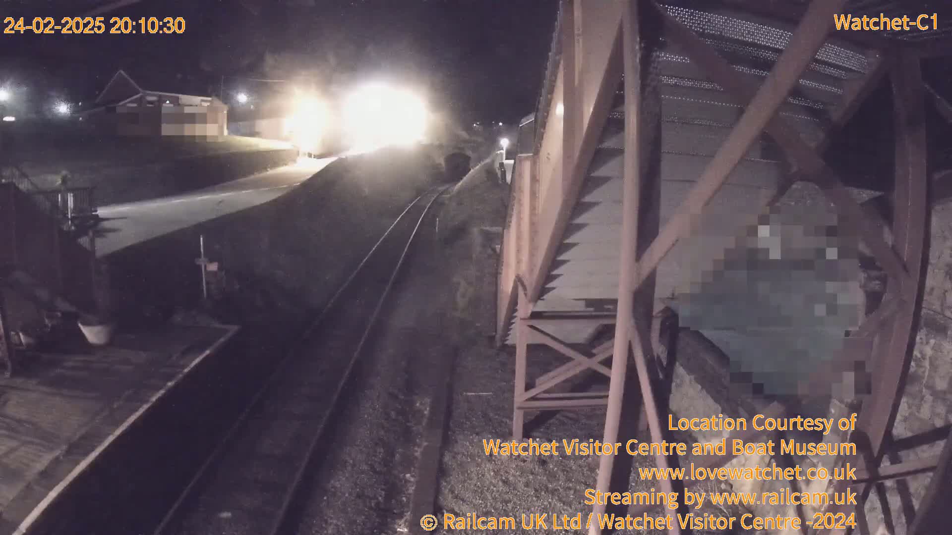 A nighttime view of a train on tracks near a building, with bright lights visible in the distance.