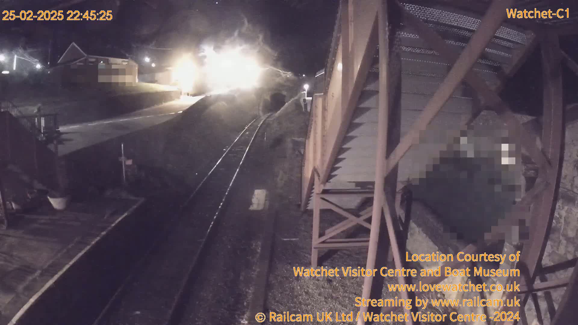 A brightly lit train passes by a railway station at night.