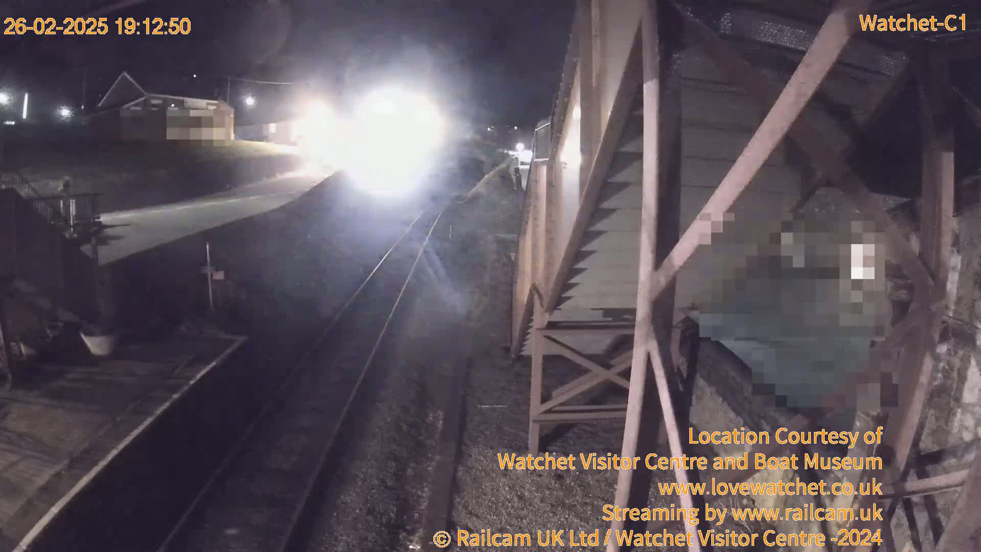 At night, a train approaches a railway station, which is partially obscured by a metal structure.
