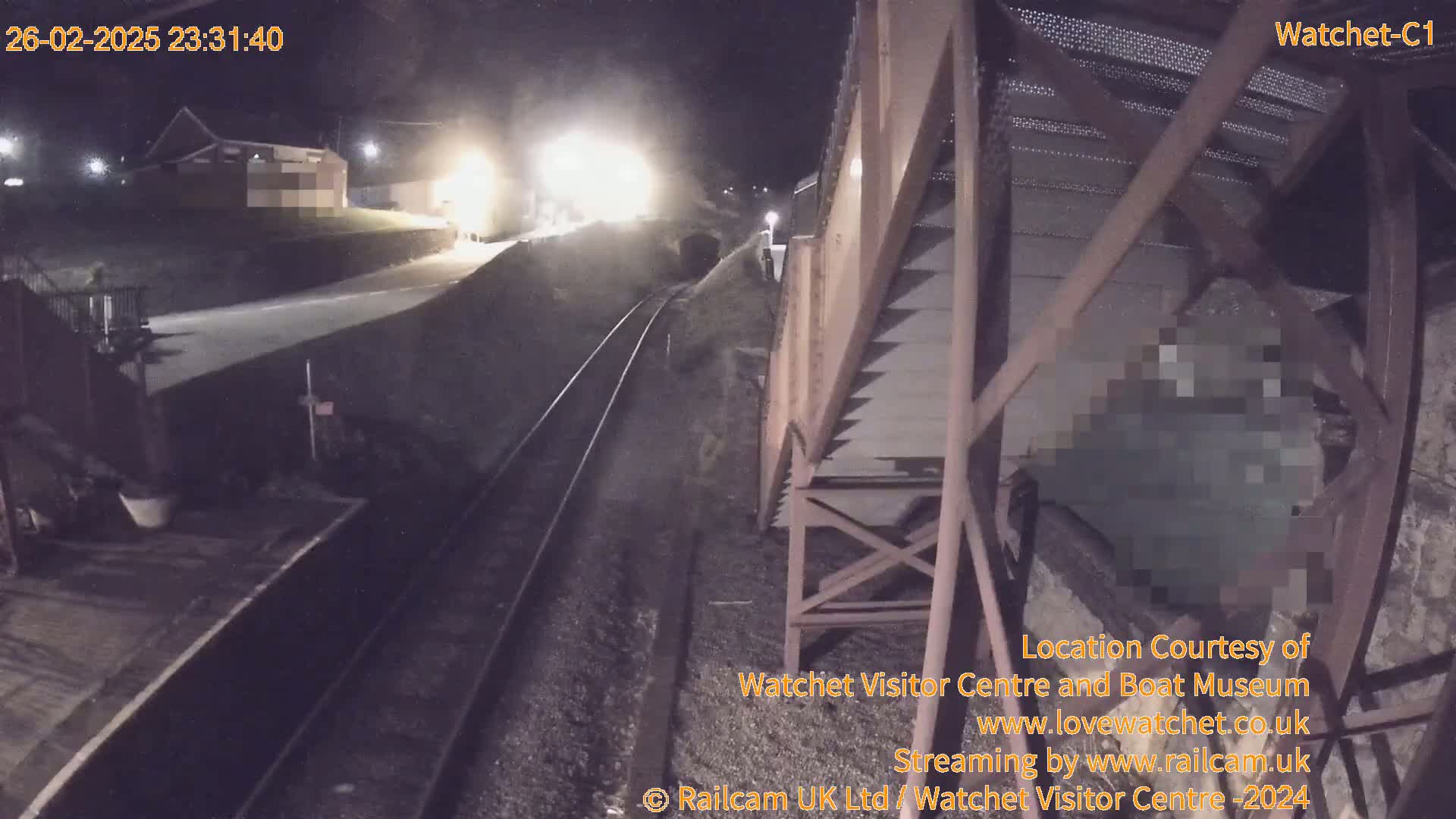 At night, a train approaches a railway station under a metal structure, with a building visible in the background.