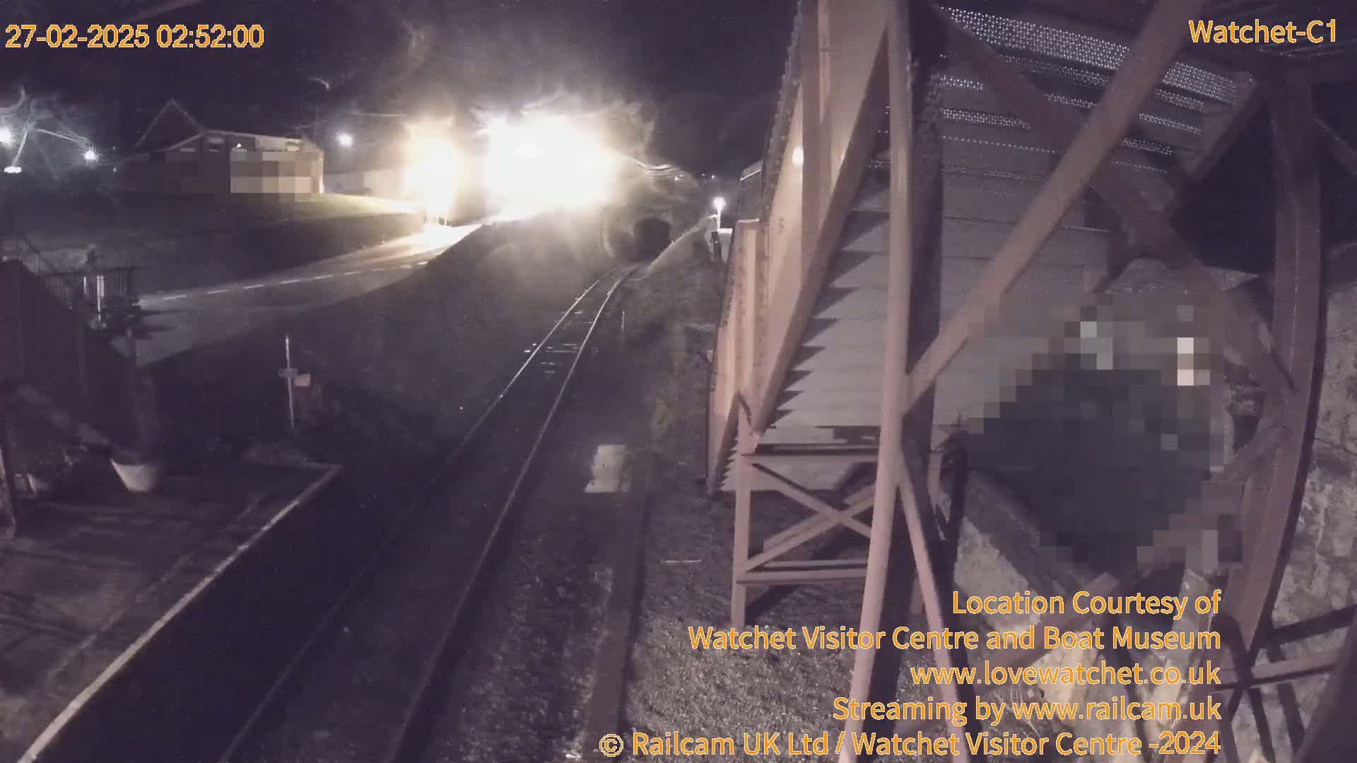 At night, a train emerges from a tunnel, its headlights illuminating the area and a nearby structure; the scene is captured by a security camera.