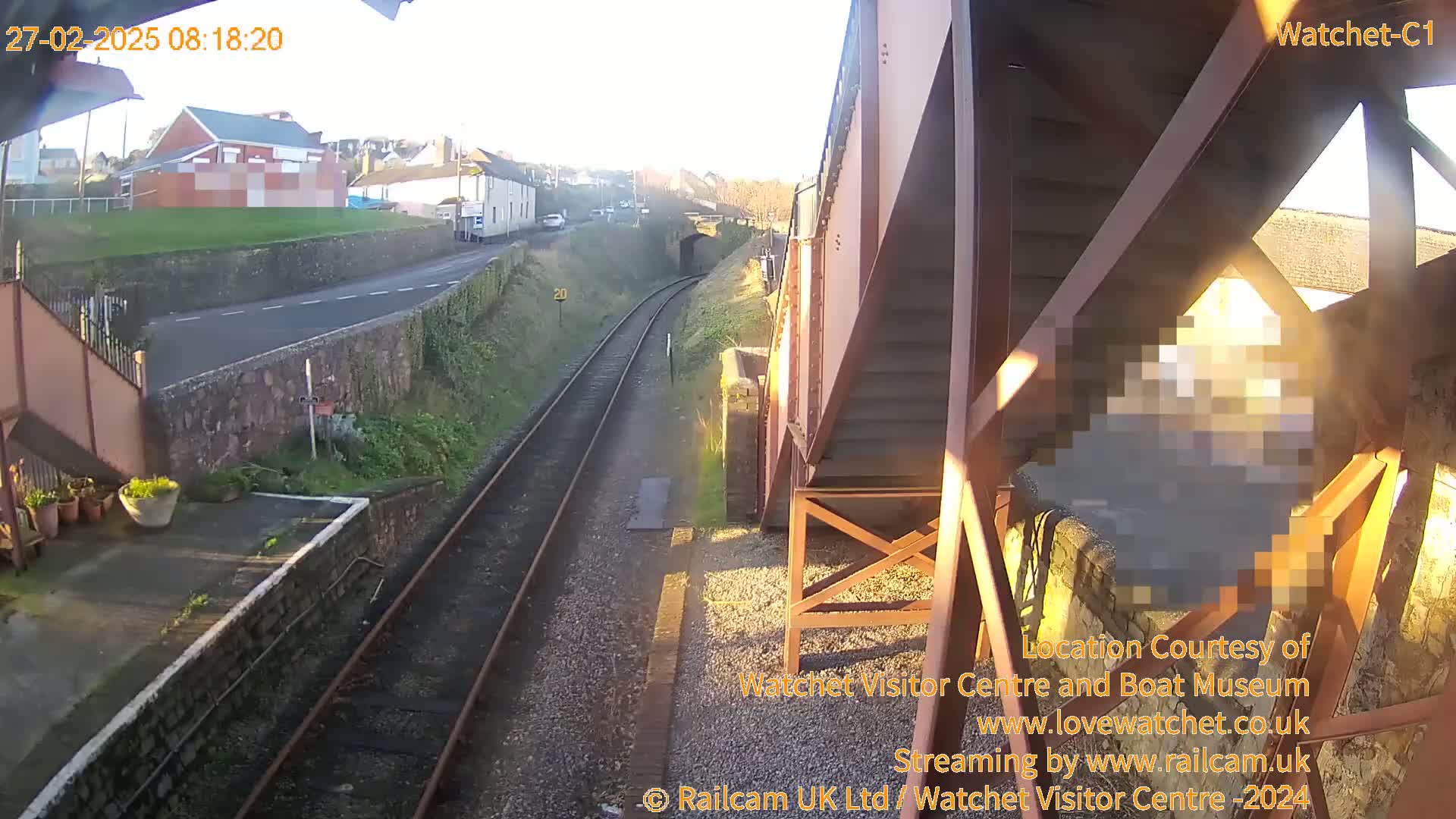 A sunny view from a train platform shows railroad tracks leading toward a tunnel, with a road and buildings visible in the background.