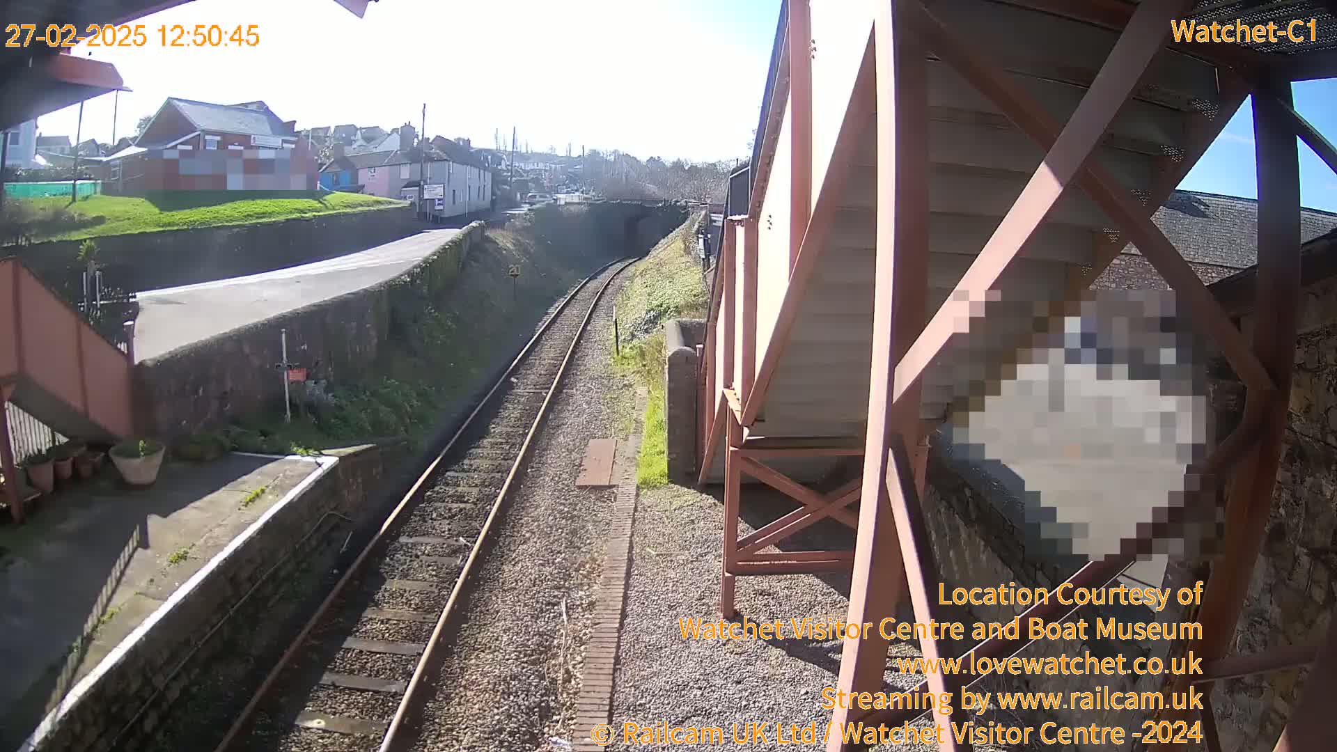 A sunny view from a railway platform shows train tracks extending into the distance, passing under a bridge and alongside buildings.