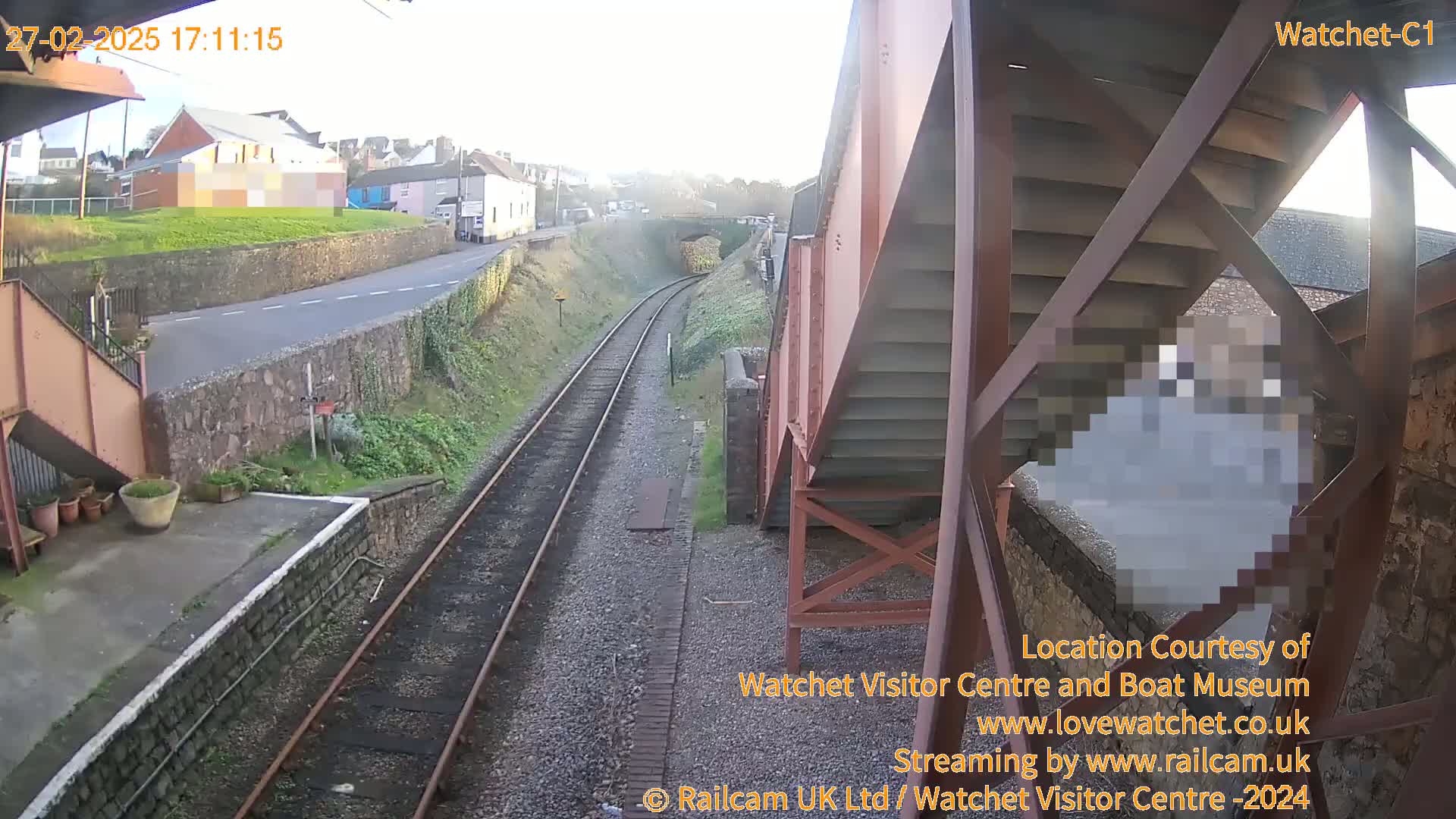 A view from a railway footbridge on a sunny day shows train tracks, a road, and buildings in the distance.