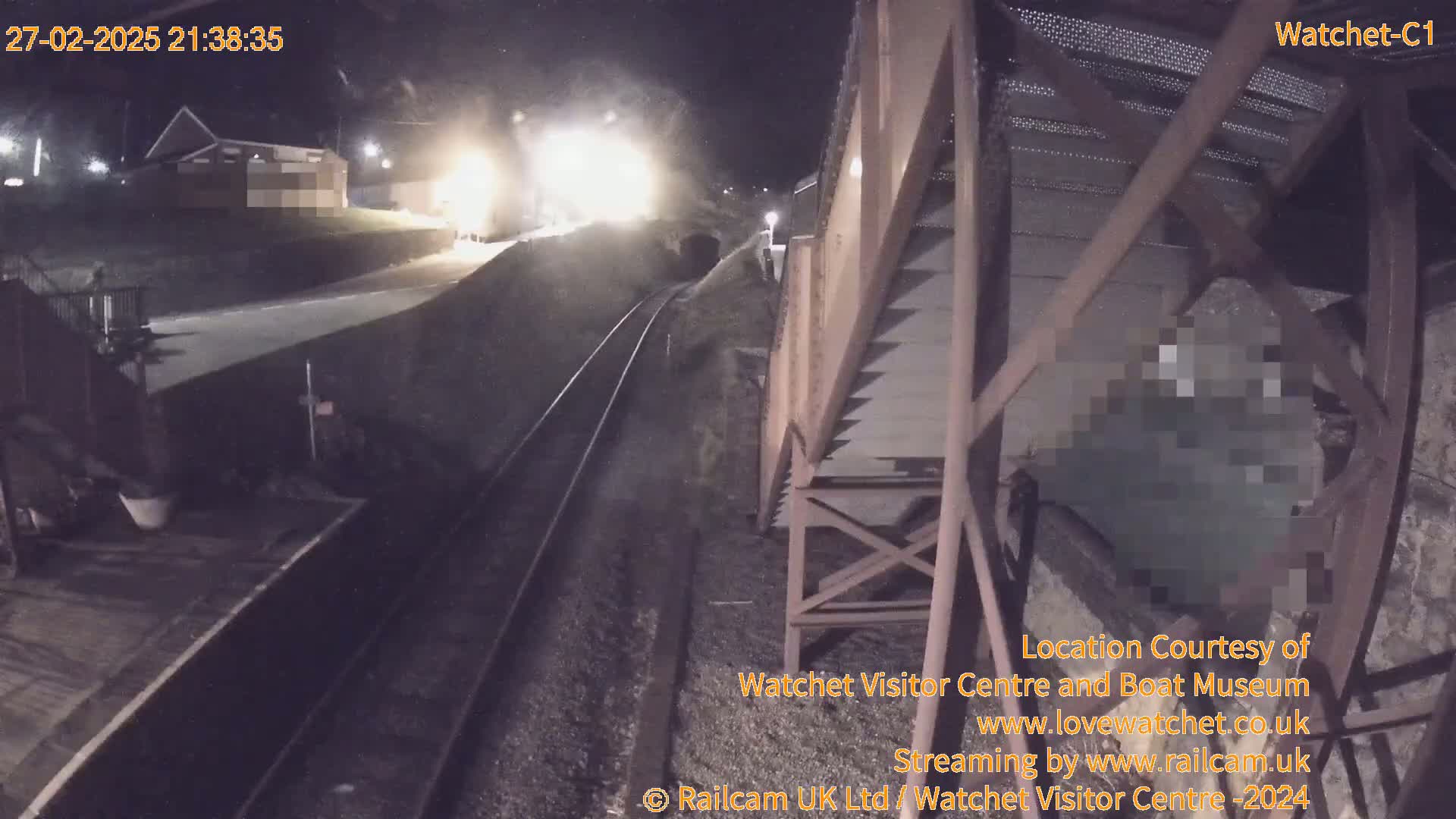 A nighttime view from a high vantage point shows a train approaching a tunnel, with a building visible in the background under dark skies.