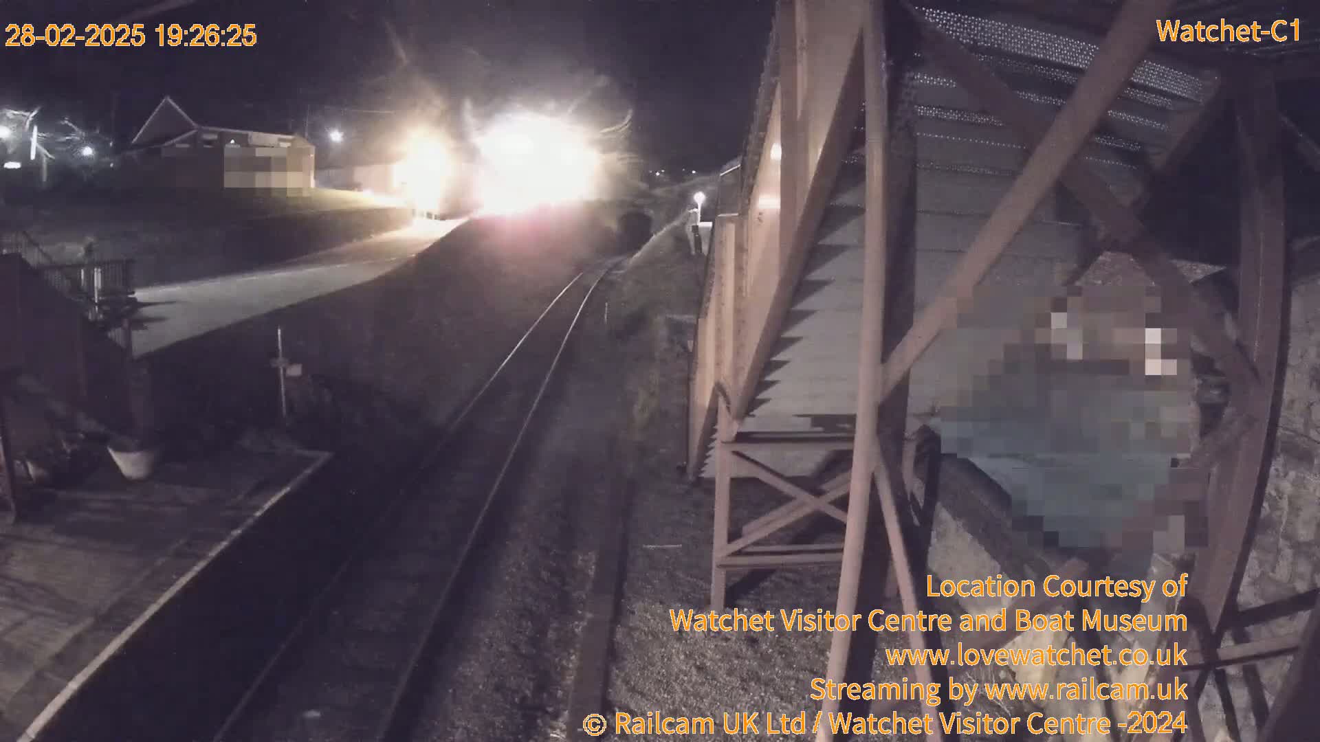 A nighttime view from an elevated vantage point shows a train approaching a station under a dark, clear sky.