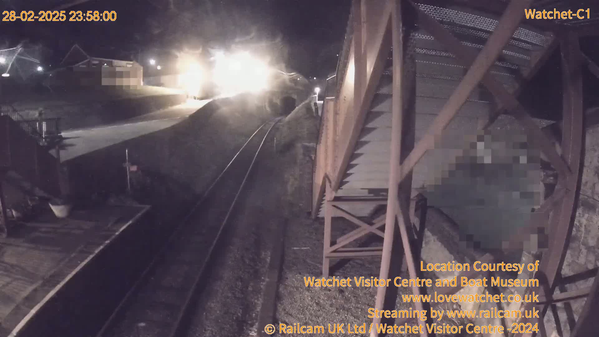 A nighttime, low-angle view shows a train moving through a tunnel, leaving a trail of light and dust, beside a metal structure and railway tracks.
