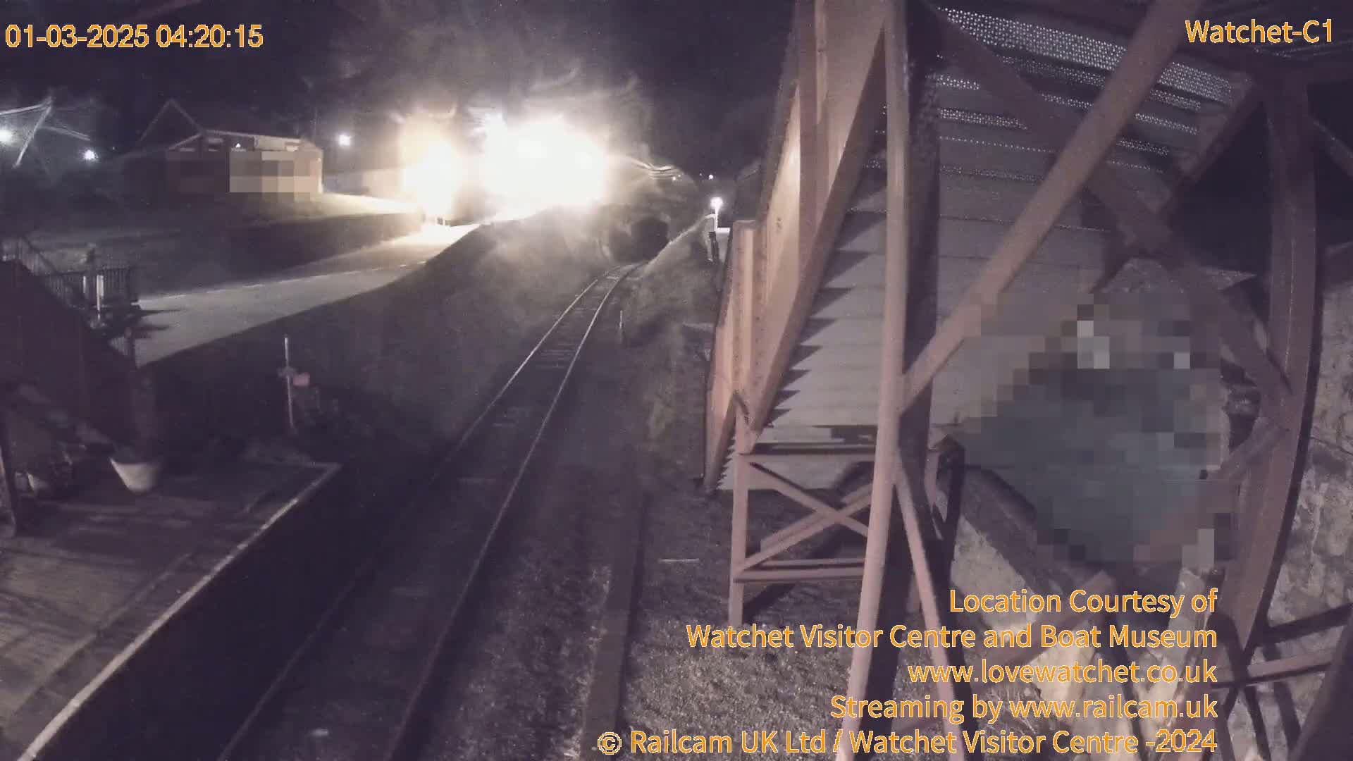 A brightly lit train emerges from a tunnel at night, passing by a railway platform and a metal structure.