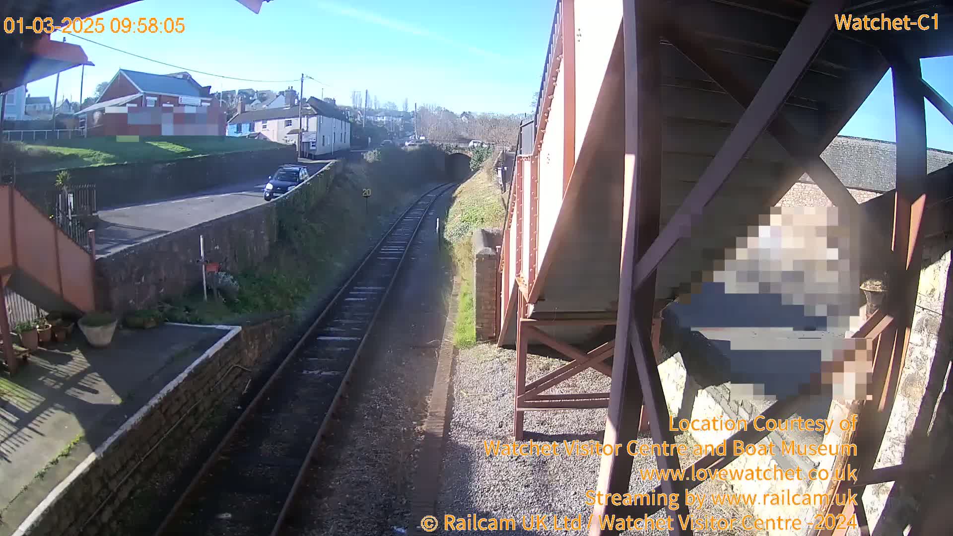 A sunny view of railroad tracks extending from a station platform, past a road with a car, toward a tunnel and residential buildings in the distance.