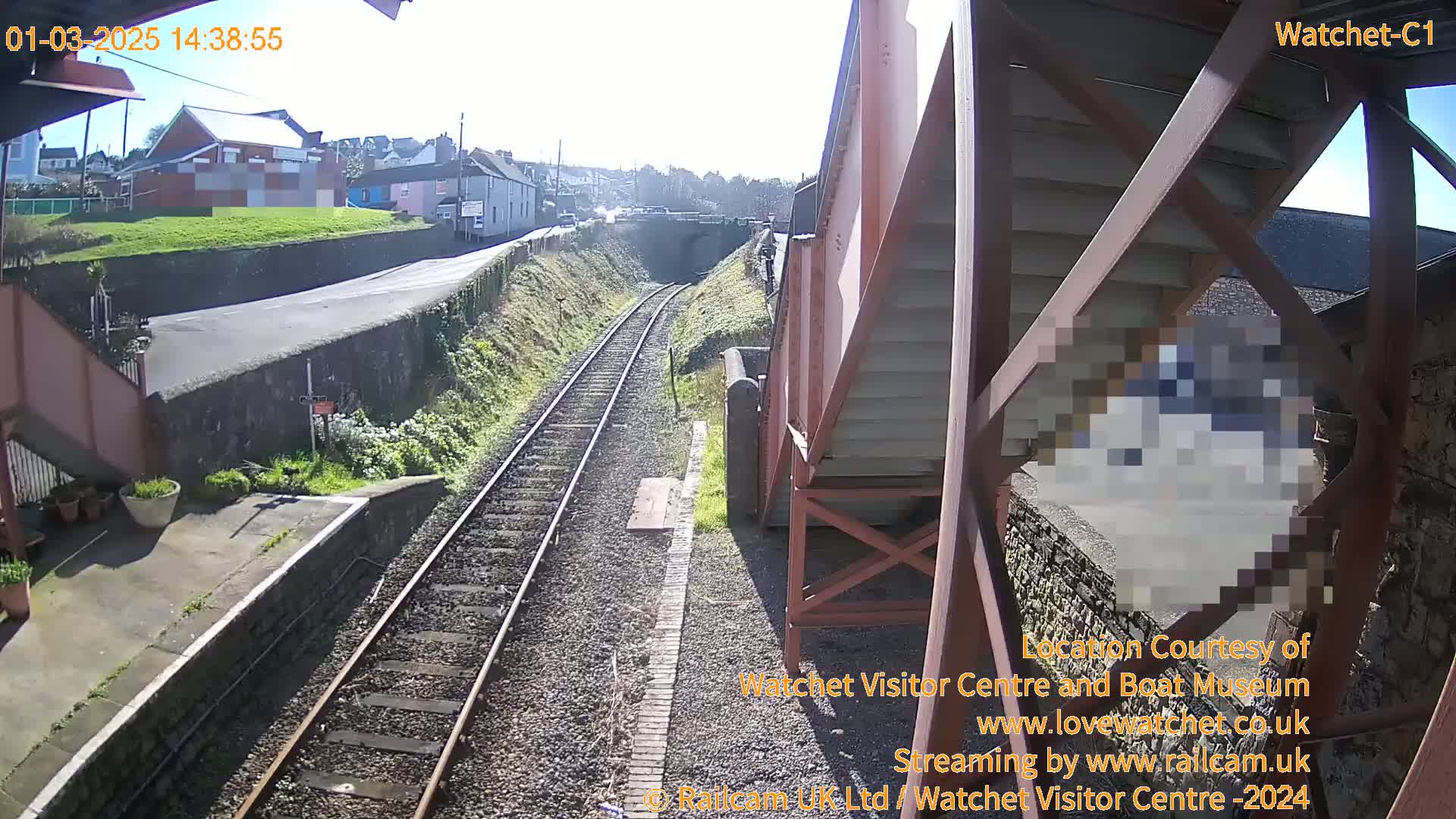 A sunny view of railway tracks and a station platform, with a road and buildings visible in the background.