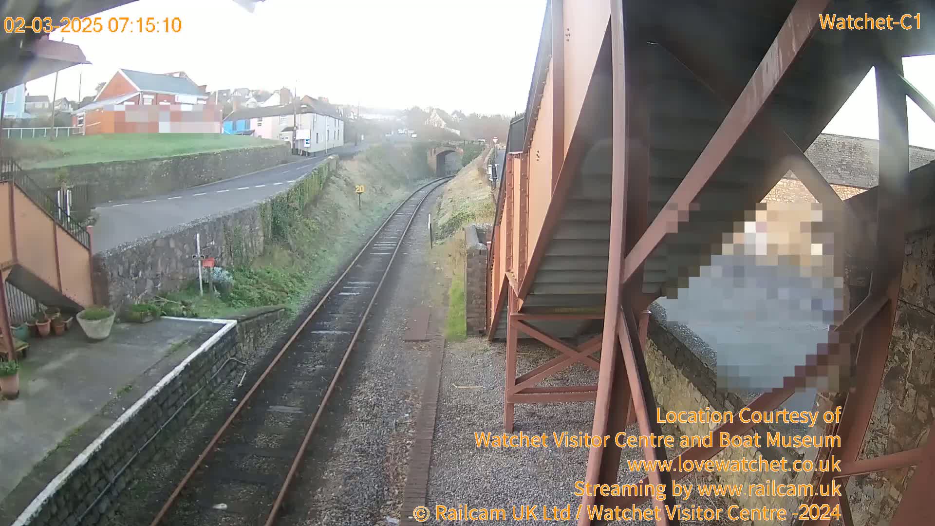 A view from a train station overpass shows a railway line extending into the distance, under a somewhat overcast sky, with a road and buildings visible in the background.