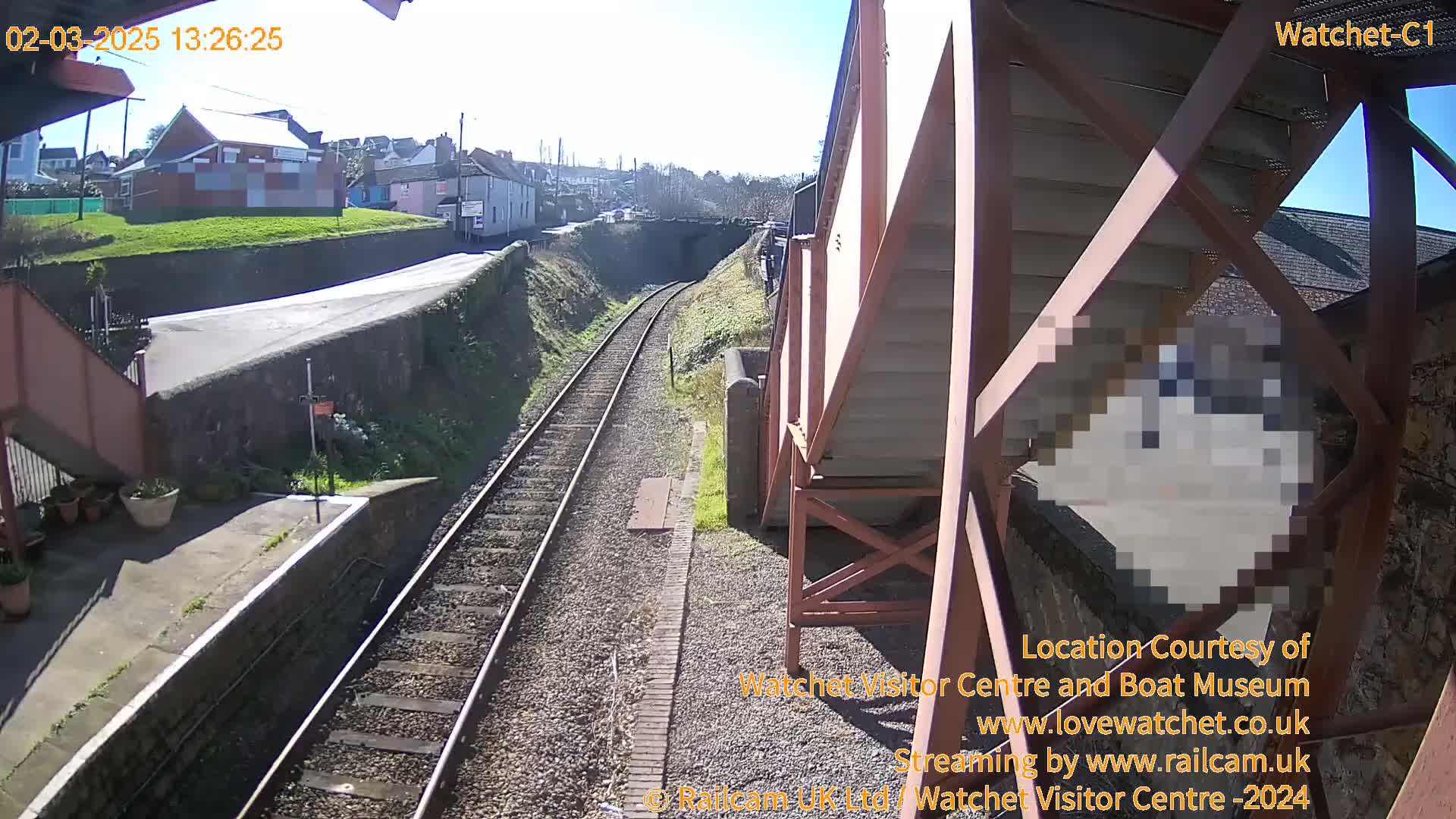 A sunny view of a railway track and station platform, with a metal structure and buildings in the background.