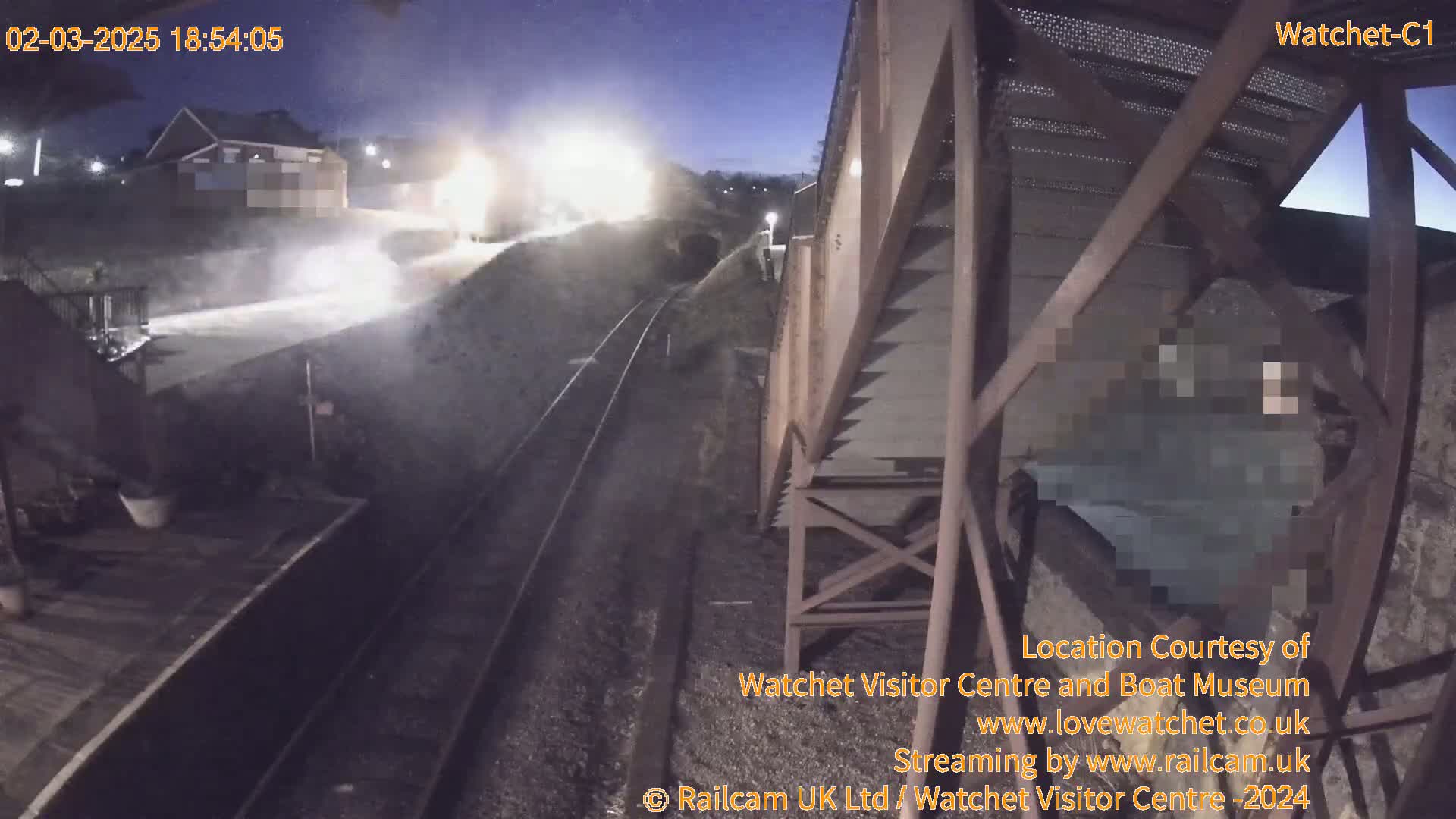 A nighttime view from a high vantage point shows a train track with a vehicle in the distance, illuminated by bright lights, under a twilight sky.