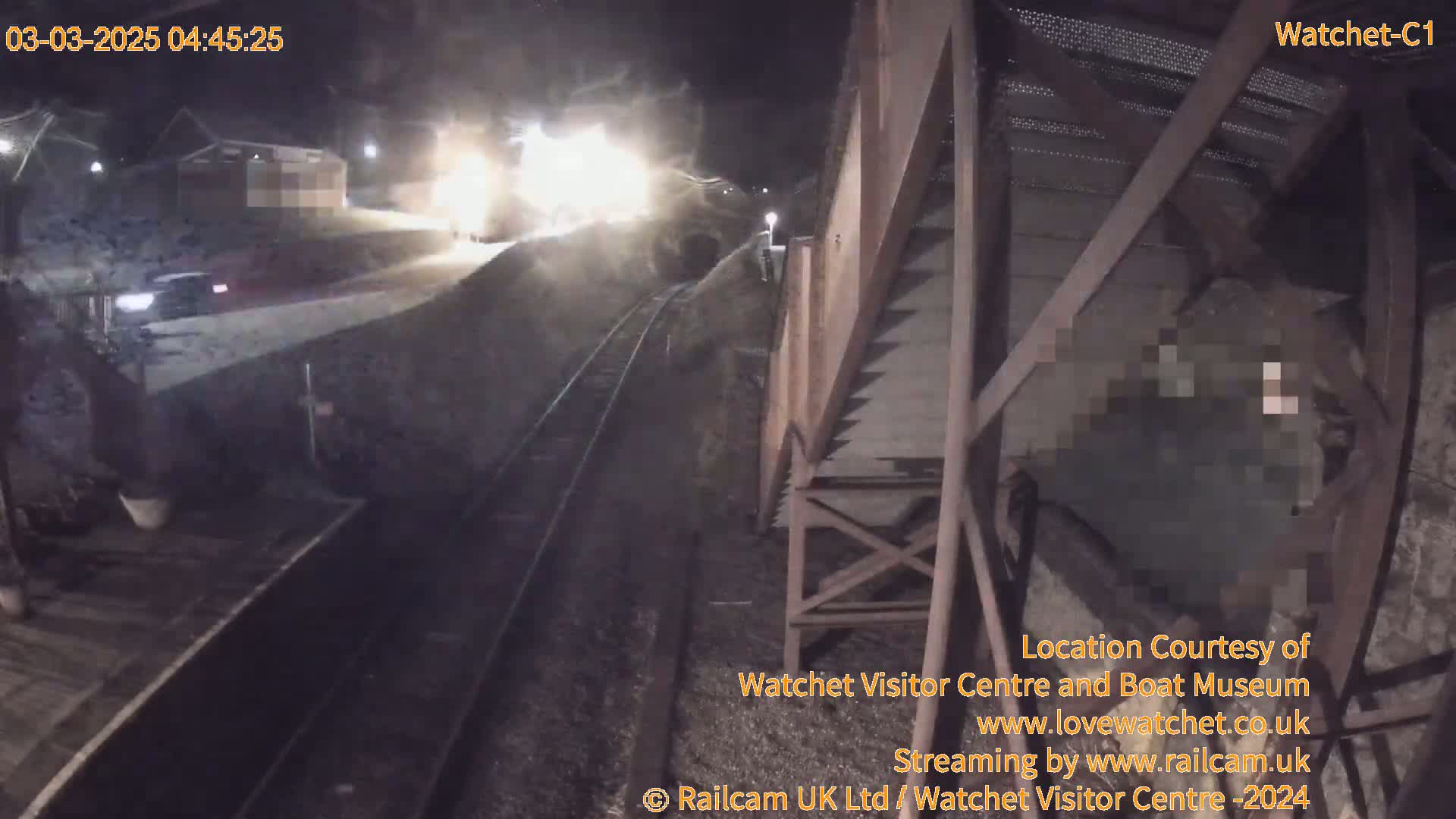 A nighttime view of a train passing by a railway station, with a vehicle parked nearby under dim lighting.