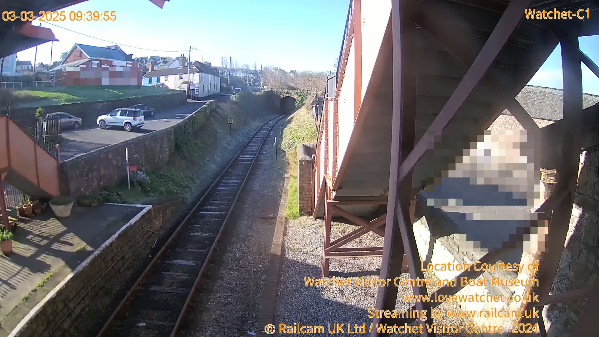 A sunny view of railway tracks extending into a tunnel, with a road and buildings visible in the background, and a few cars parked nearby.