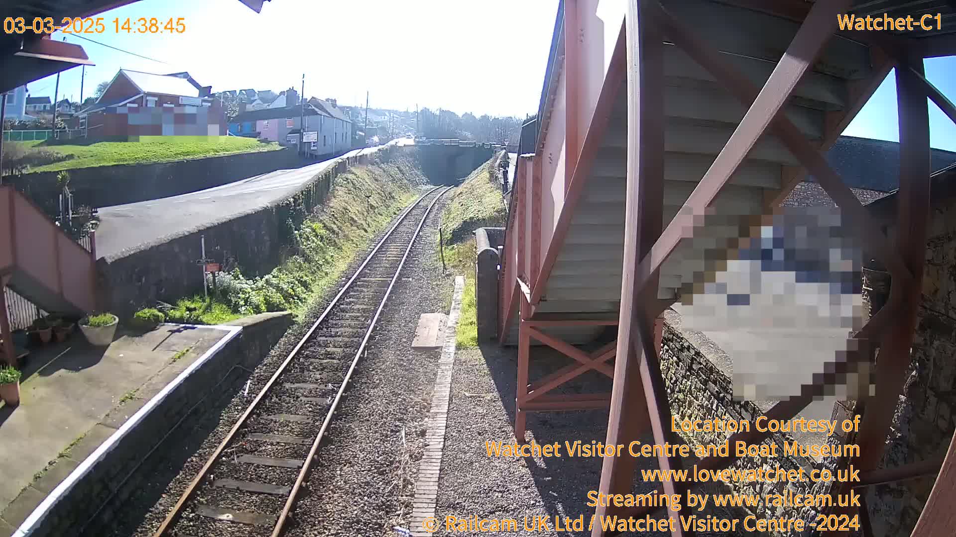 A sunny view of train tracks extending into the distance, alongside a road and buildings, seen from an elevated vantage point.