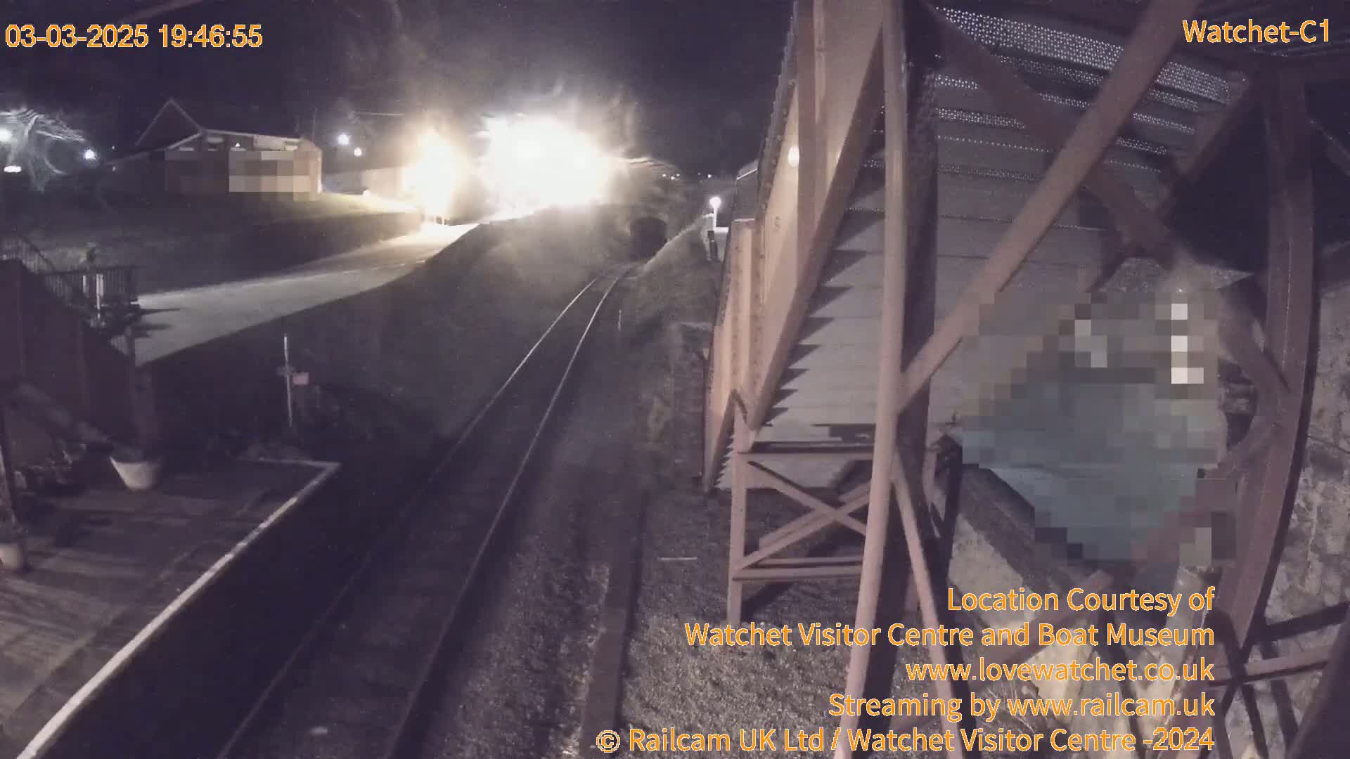 A nighttime view from a high vantage point shows a train approaching a station under bright lights, alongside railway tracks and a metal structure.
