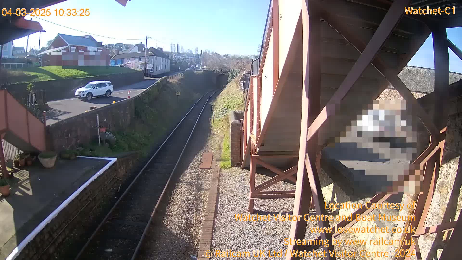 A sunny day at a railway station, viewed from a footbridge, shows a white SUV parked on a road next to the tracks.
