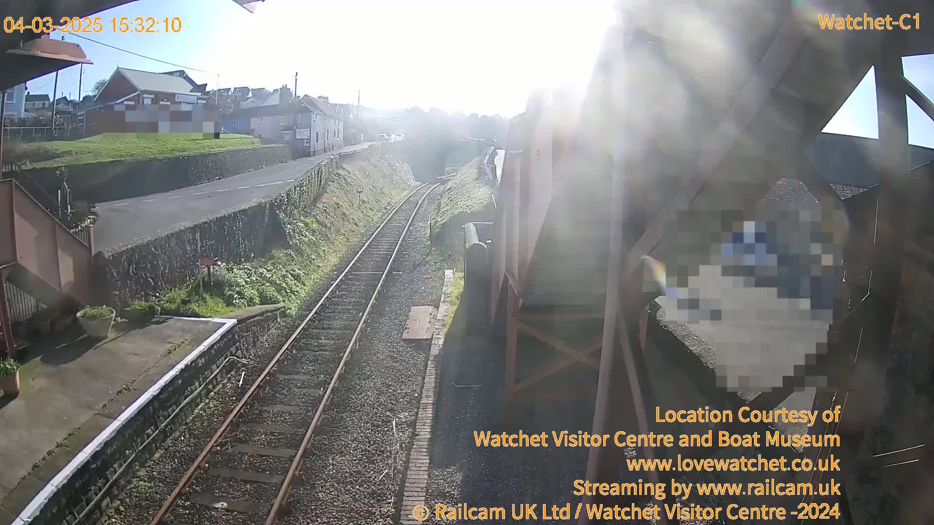 A sunny view from a train station shows train tracks leading to a tunnel, with buildings and a road visible in the background.