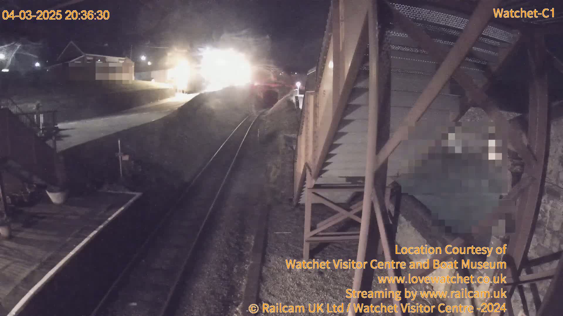 At night, a brightly lit train approaches a railway station on a track bordered by gravel, seen from a high vantage point overlooking a metal structure.