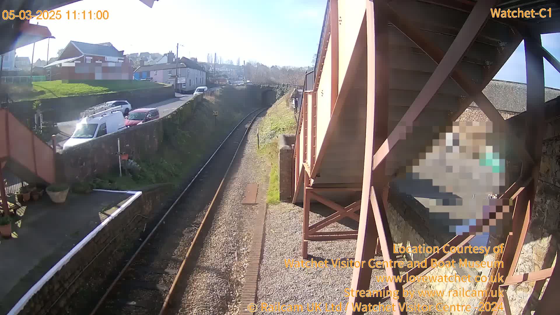 A sunny view from a railway bridge shows train tracks, a road with several cars, and a small town in the distance.