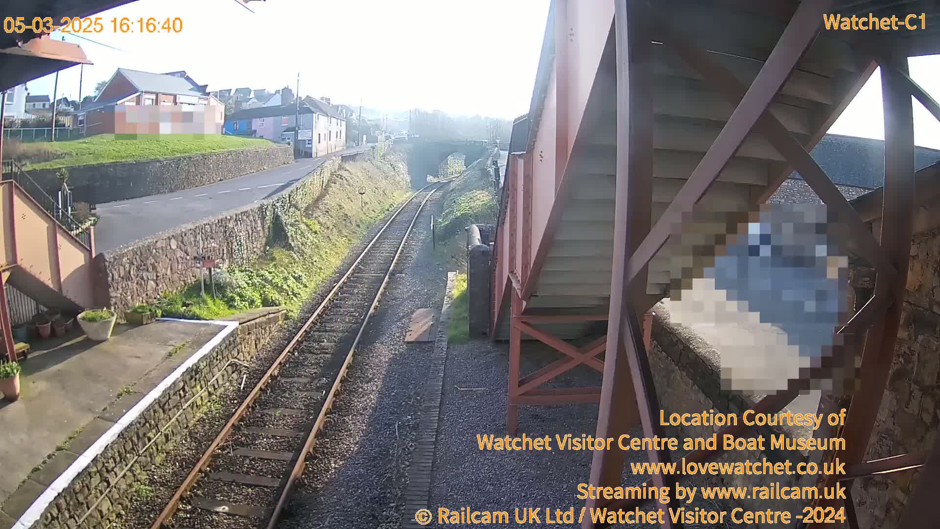 A sunny view of train tracks and a station platform, with a metal staircase and buildings in the background.