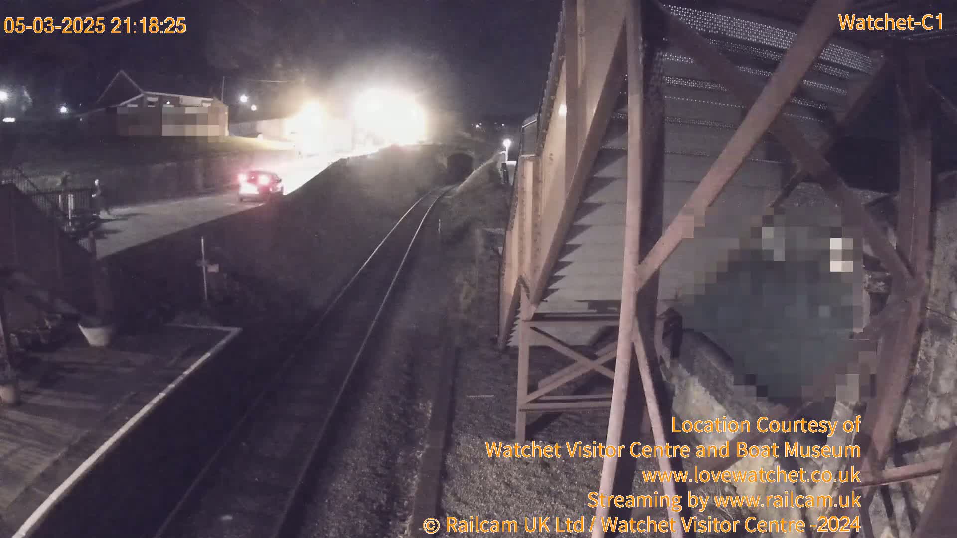 A nighttime view shows a car driving on a road next to train tracks and a large metal structure, under dark, possibly overcast conditions.