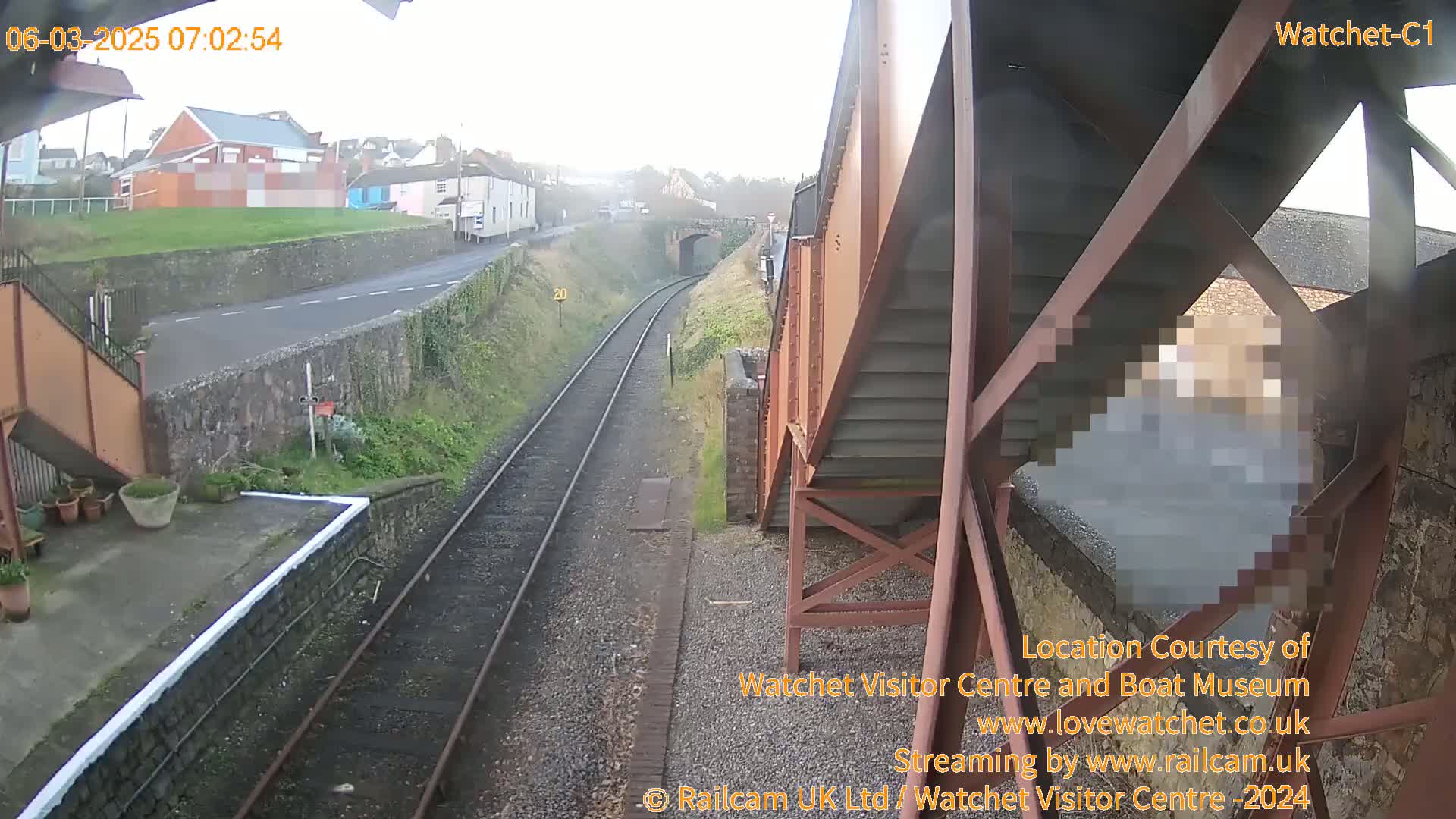 A view of a train track and a road from a slightly elevated position on an overcast day, showing a partially obscured person on a metal staircase structure.