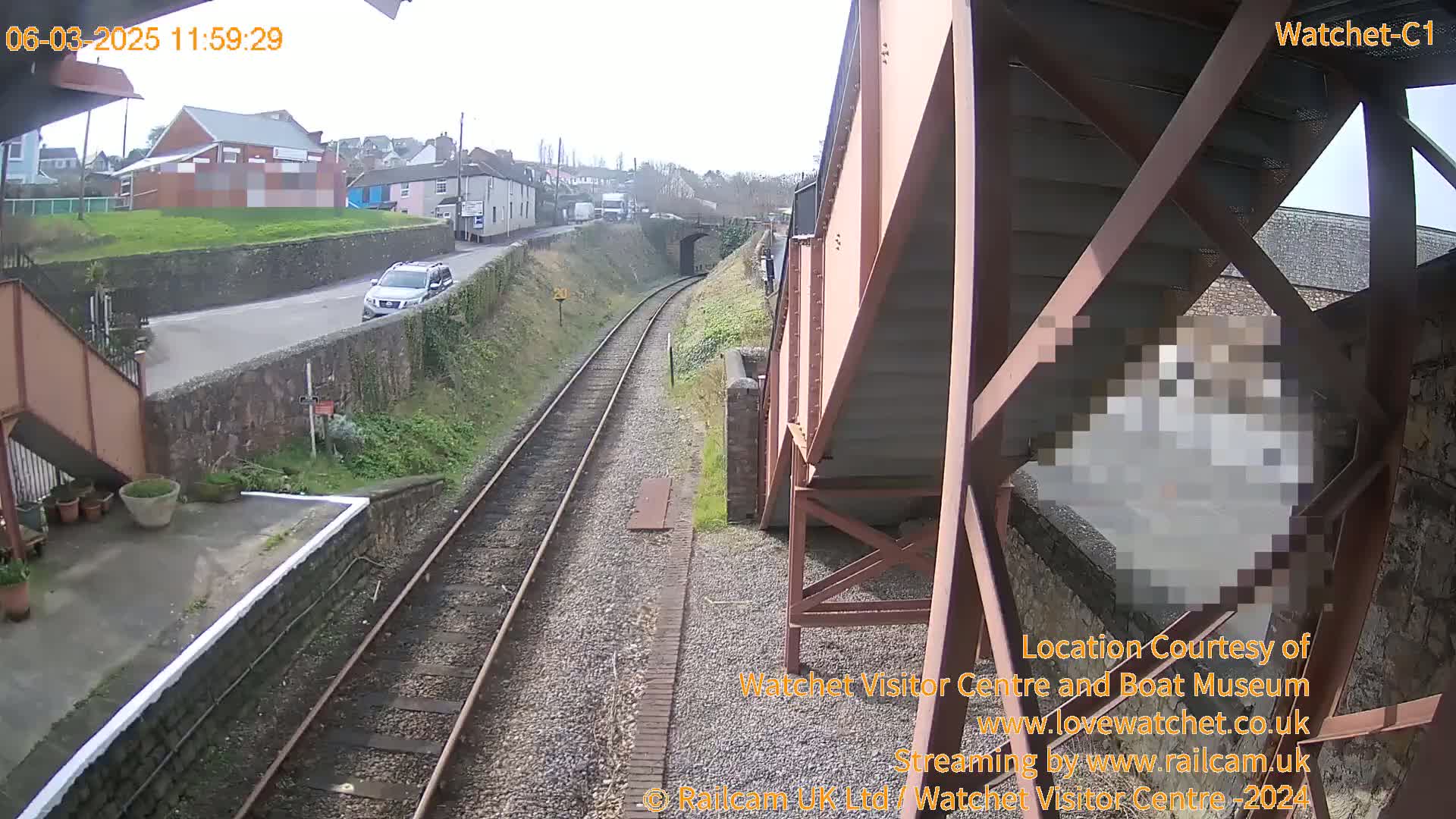 A view from a railway bridge on an overcast day shows train tracks, a road with a car, and buildings in the distance.