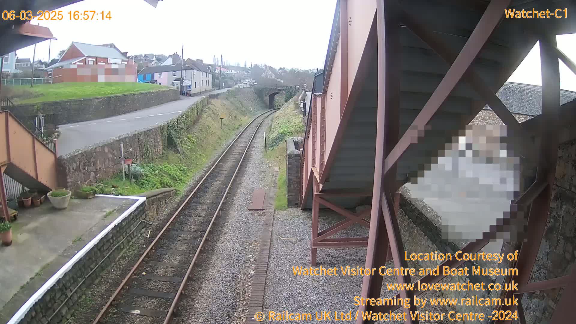 A view from a railway bridge on an overcast day shows train tracks leading toward a tunnel and a road passing by buildings.