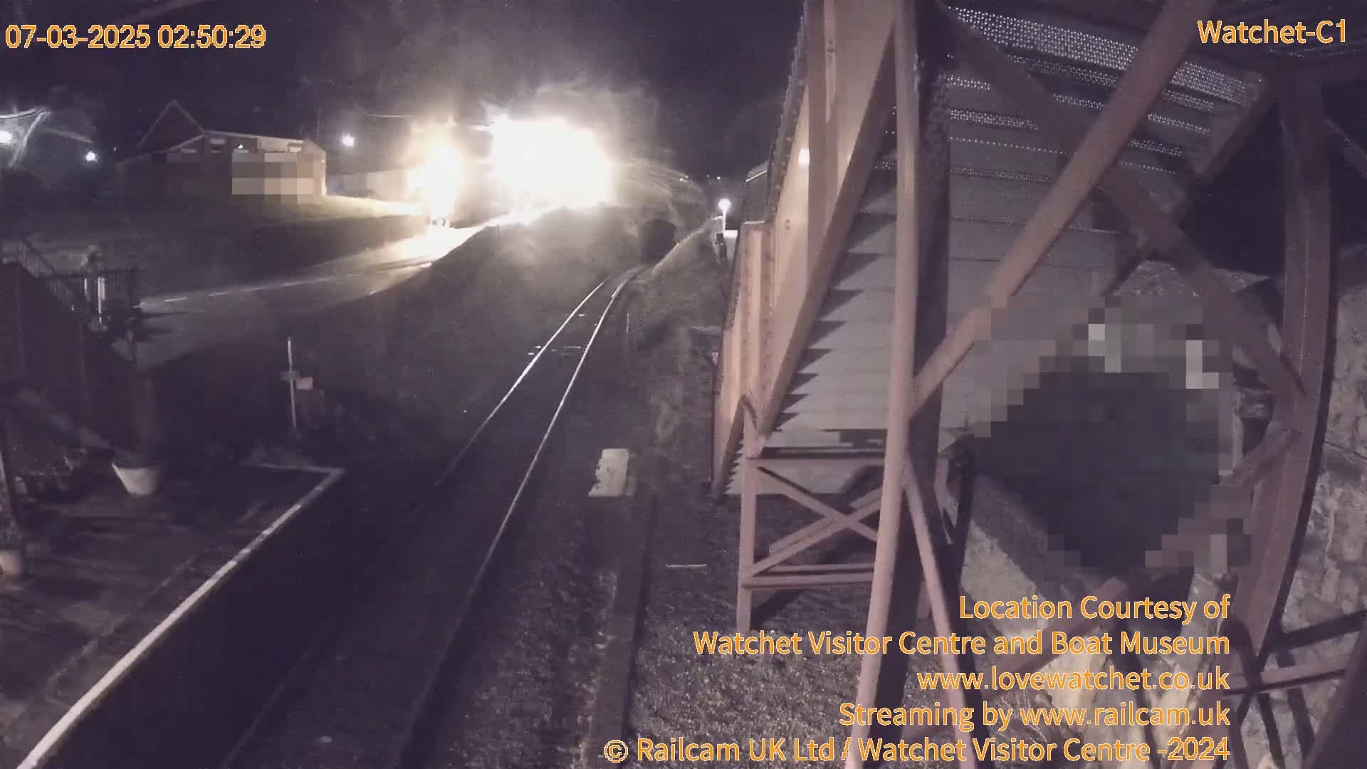 A nighttime, low-light view shows a train approaching a tunnel from the perspective of a metal structure beside the tracks.