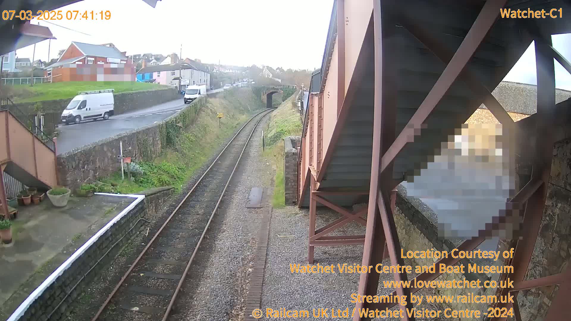 A view from under a railway bridge on an overcast day shows railway tracks leading to a tunnel, with a road and two white vans visible in the distance.
