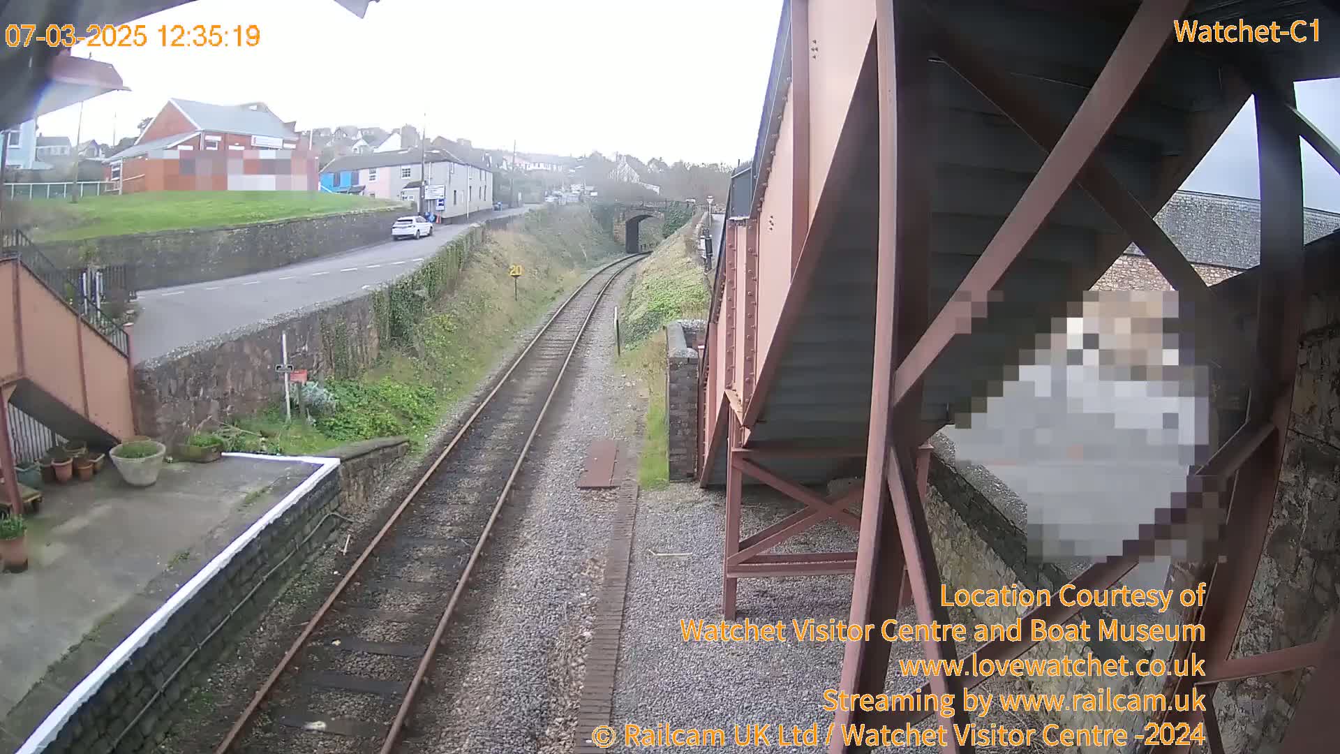 A view from a railway bridge on an overcast day shows train tracks leading to a town with buildings and a road, and a person is partially visible under the bridge.