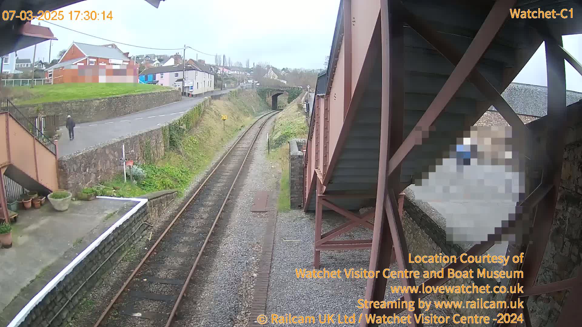 A view from a railway bridge on an overcast day shows train tracks, a person walking on a road, and buildings in the distance.