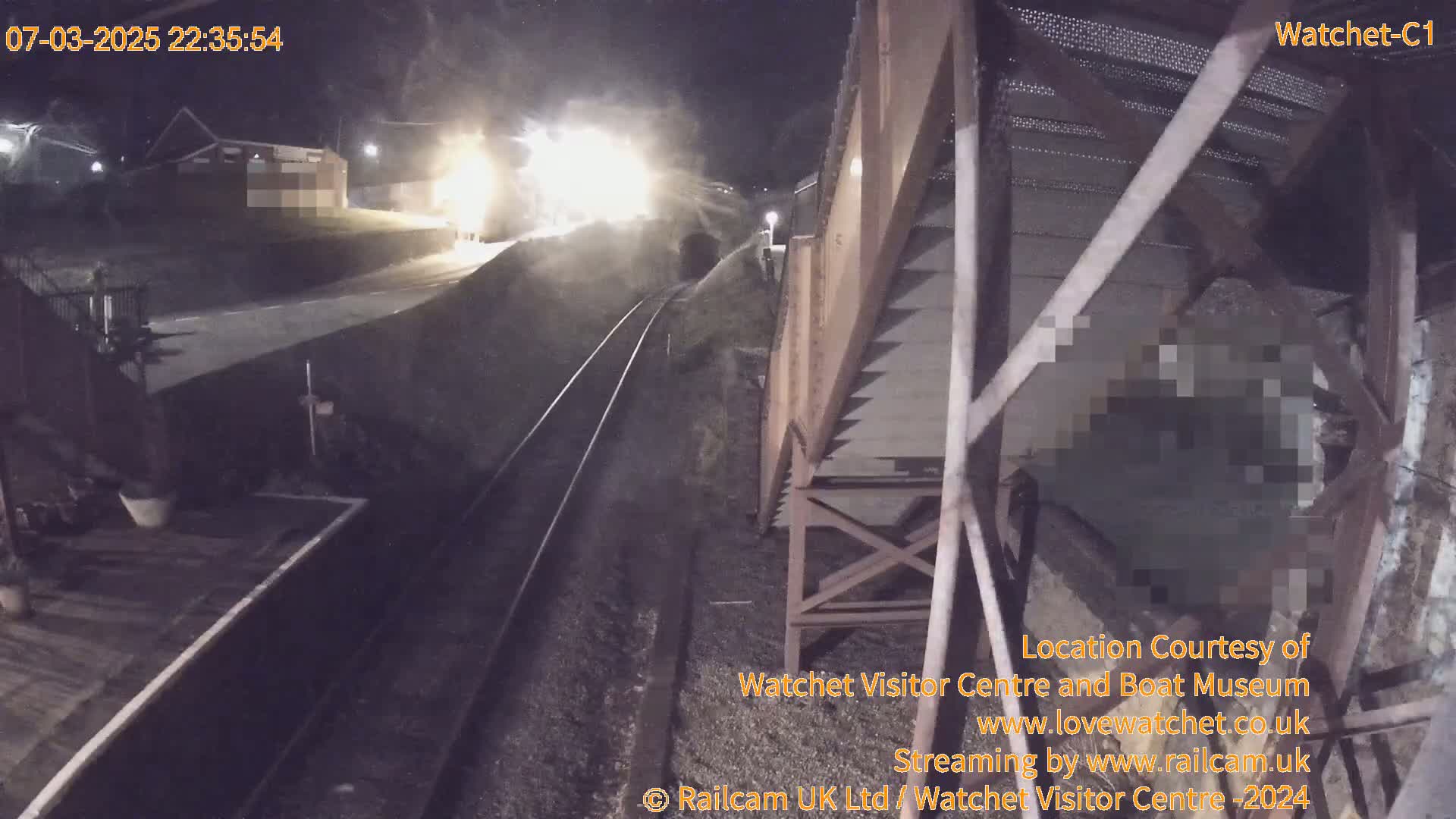 A nighttime view from a train car shows a brightly lit train approaching a tunnel, alongside railroad tracks and a dark embankment.