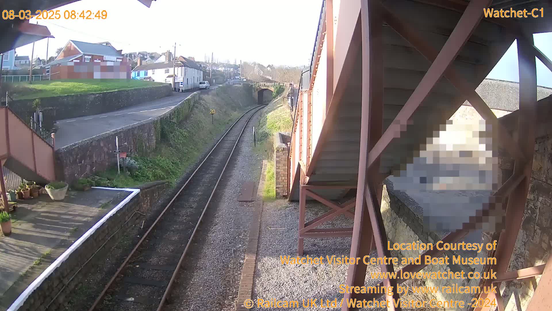 A sunny view of a railway track and a station platform from under a metal footbridge, with a road and buildings visible in the background.