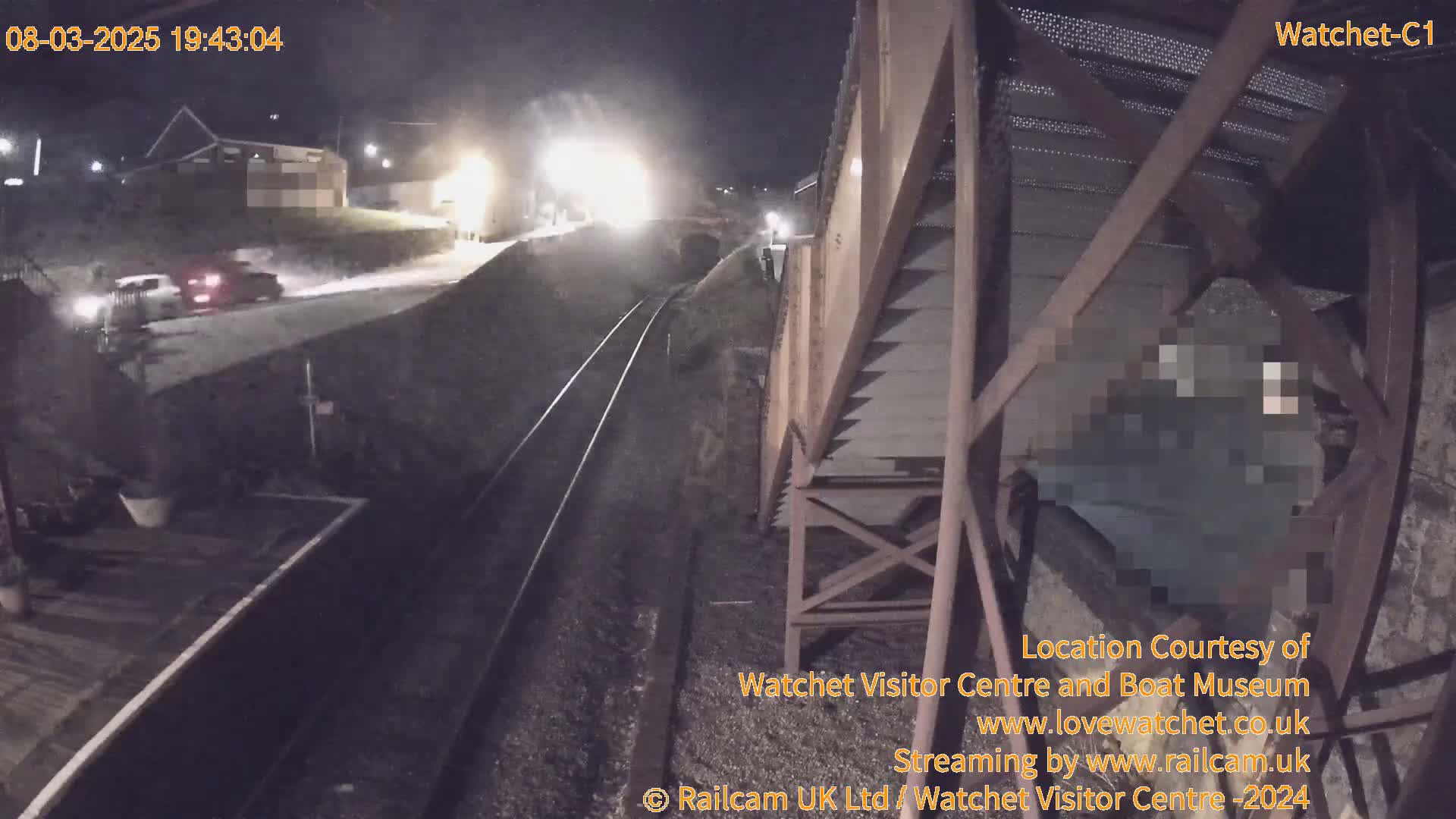 A nighttime view from a bridge shows a roadway with two vehicles and train tracks below, under dark, possibly overcast conditions.