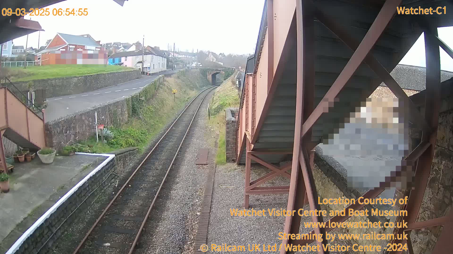 A view from a railway bridge on an overcast day shows train tracks leading to a road and buildings in the distance.